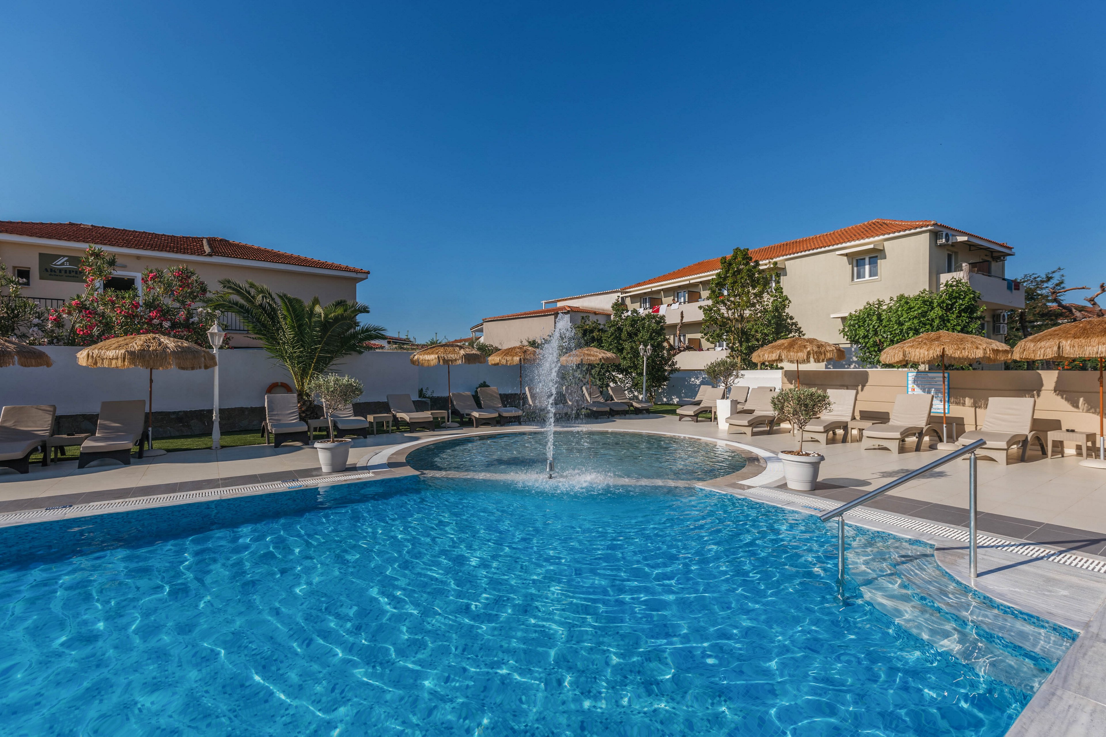 A bright blue swimming pool with a central fountain sits in the foreground of a sunny resort. Lounge chairs and straw parasols line the poolside area, creating a relaxing vacation atmosphere. Low-rise buildings with red-tiled roofs and lush greenery surround the pool. The clear sky and calm water emphasize a warm, tranquil holiday setting.