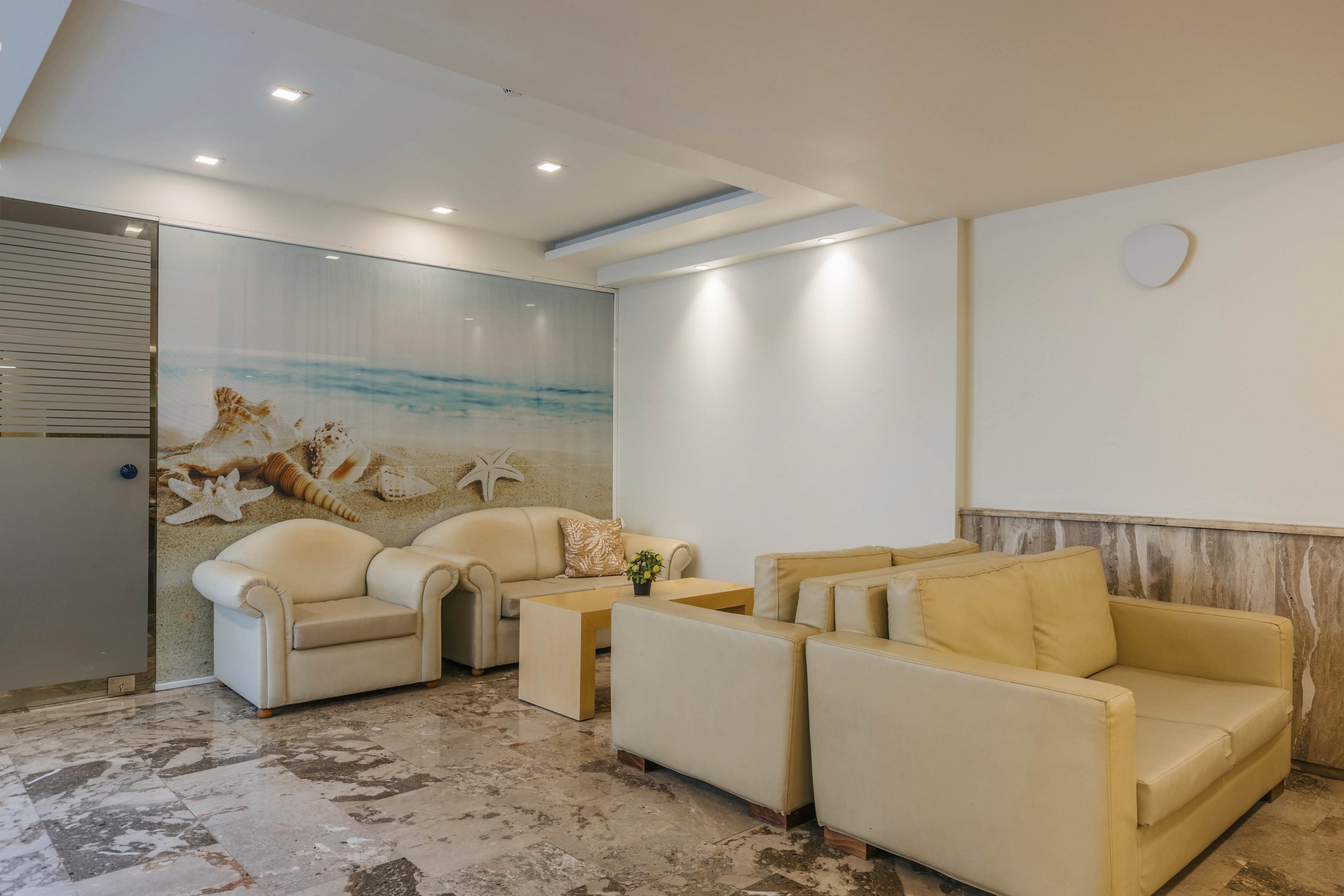 A contemporary lounge area features cream-colored sofas arranged around a small wooden coffee table. The space has a polished stone floor and neutral walls, creating a clean and minimalist look. A large wall mural depicting a beach scene with seashells and starfish adds a coastal accent to the room. Recessed ceiling lights and wall-mounted fixtures provide soft, even illumination, enhancing the calm and welcoming atmosphere.