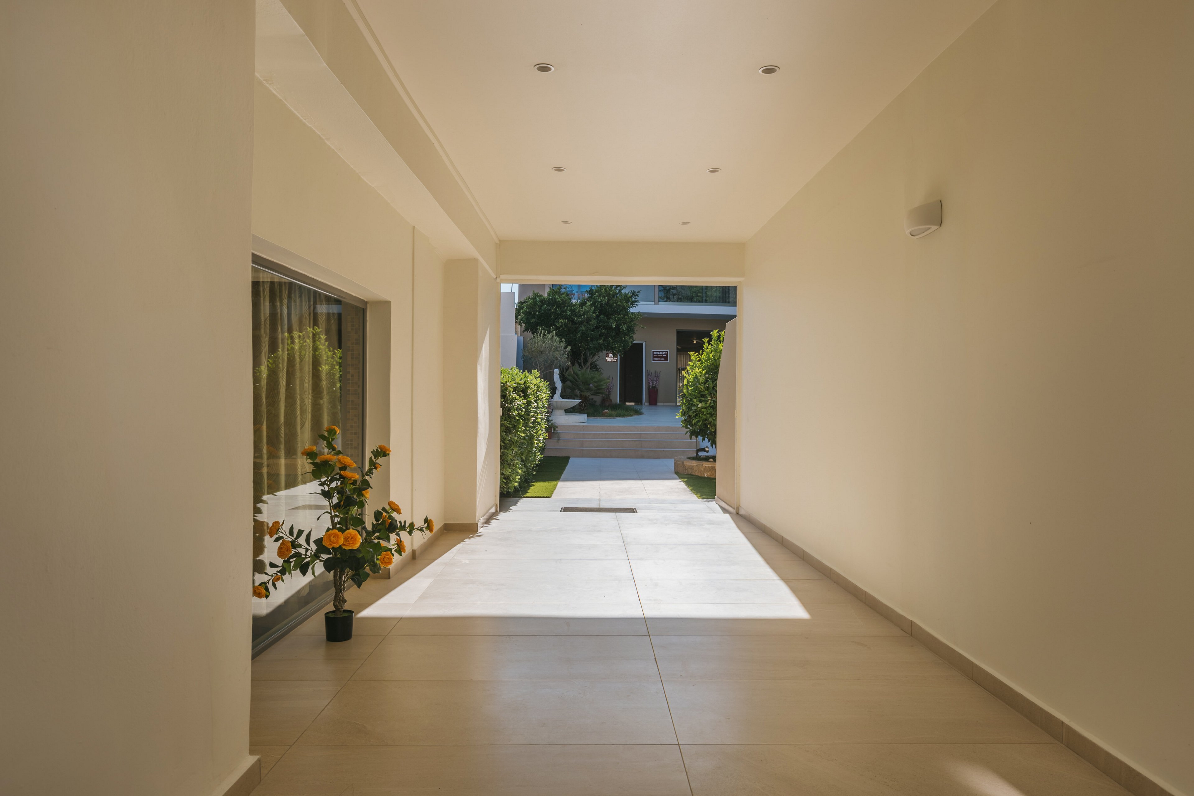 A bright, minimalist corridor opens toward a small garden and residential entrance. Smooth light-colored tiles and clean white walls create a modern, airy feel. Natural light floods the passageway, casting soft shadows on the floor. A potted flowering plant adds a touch of color near a large window on the left side.