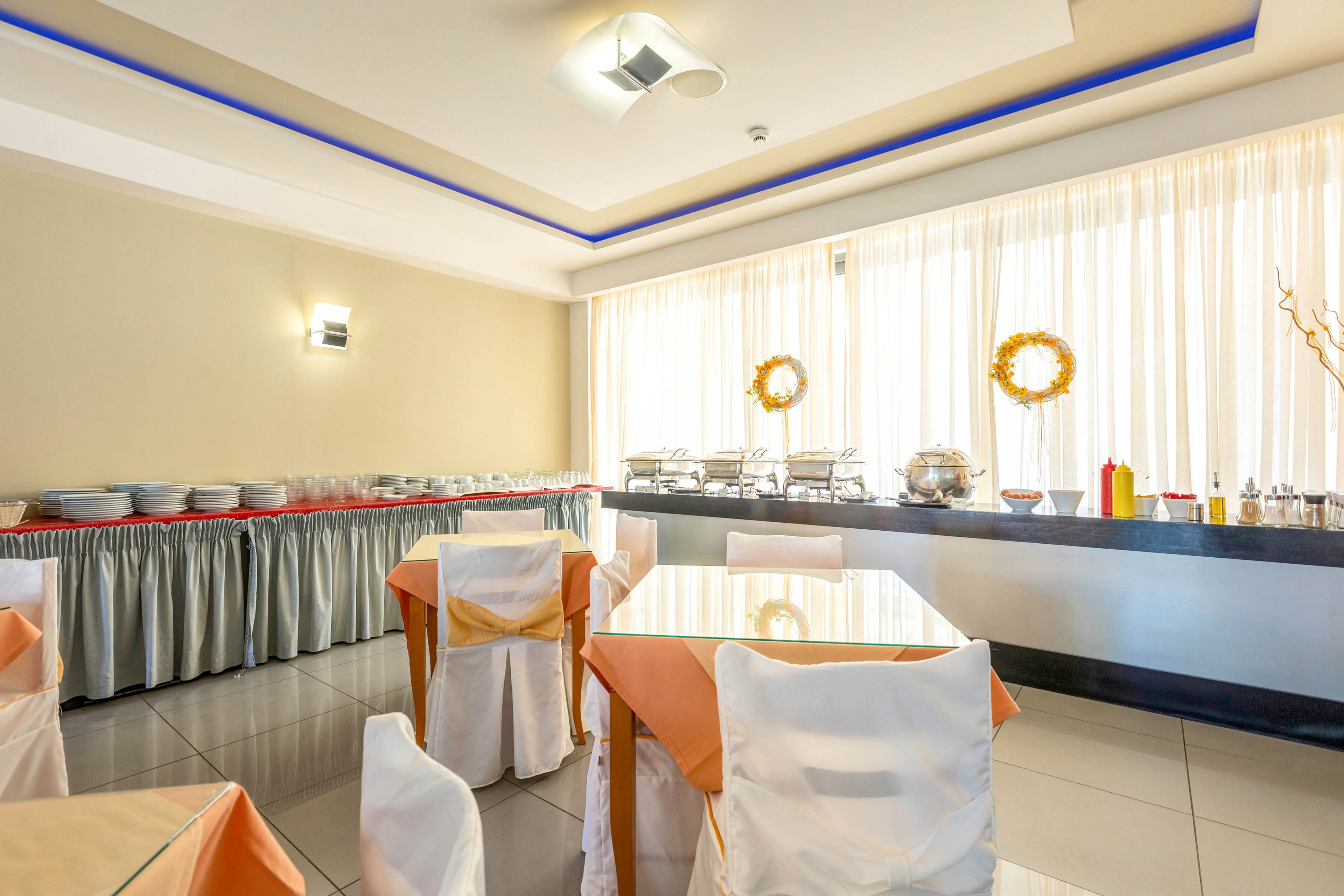 A modern buffet dining room is set up with neatly arranged tables covered in orange tablecloths and white chair covers. Along the wall, a long buffet counter holds chafing dishes and serving utensils ready for service. Large windows with sheer curtains allow natural light to fill the space, creating a bright and airy atmosphere. Decorative wreaths and minimalist lighting add a subtle, contemporary touch to the room.