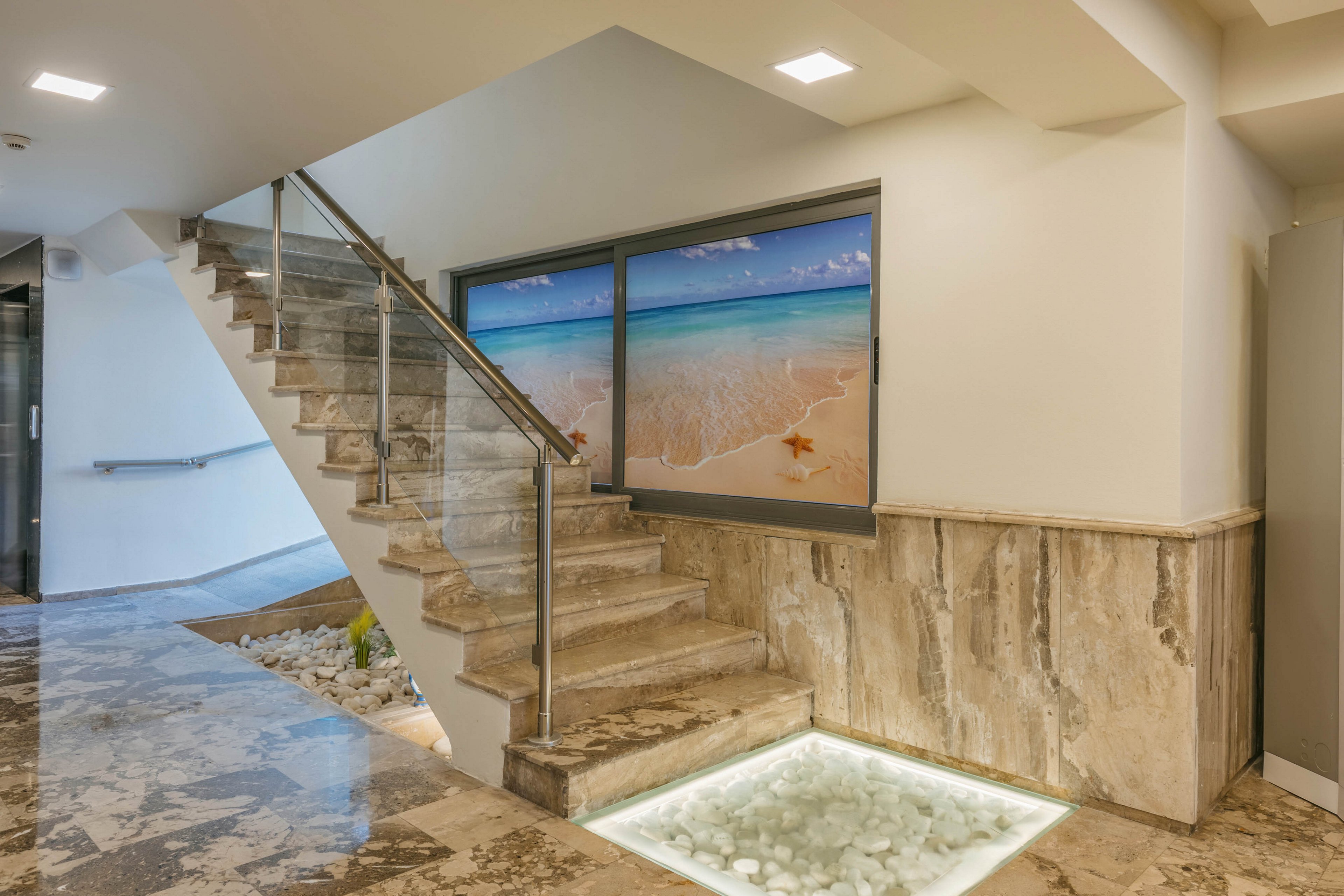 A contemporary indoor staircase with marble steps and glass railing is featured in this image. The stairwell is decorated with a large wall artwork depicting a tropical beach with turquoise water and starfish. A lit glass floor section filled with white stones adds a decorative accent at the base of the stairs. The space has polished stone flooring, neutral walls, and recessed ceiling lighting, creating a clean and modern atmosphere.