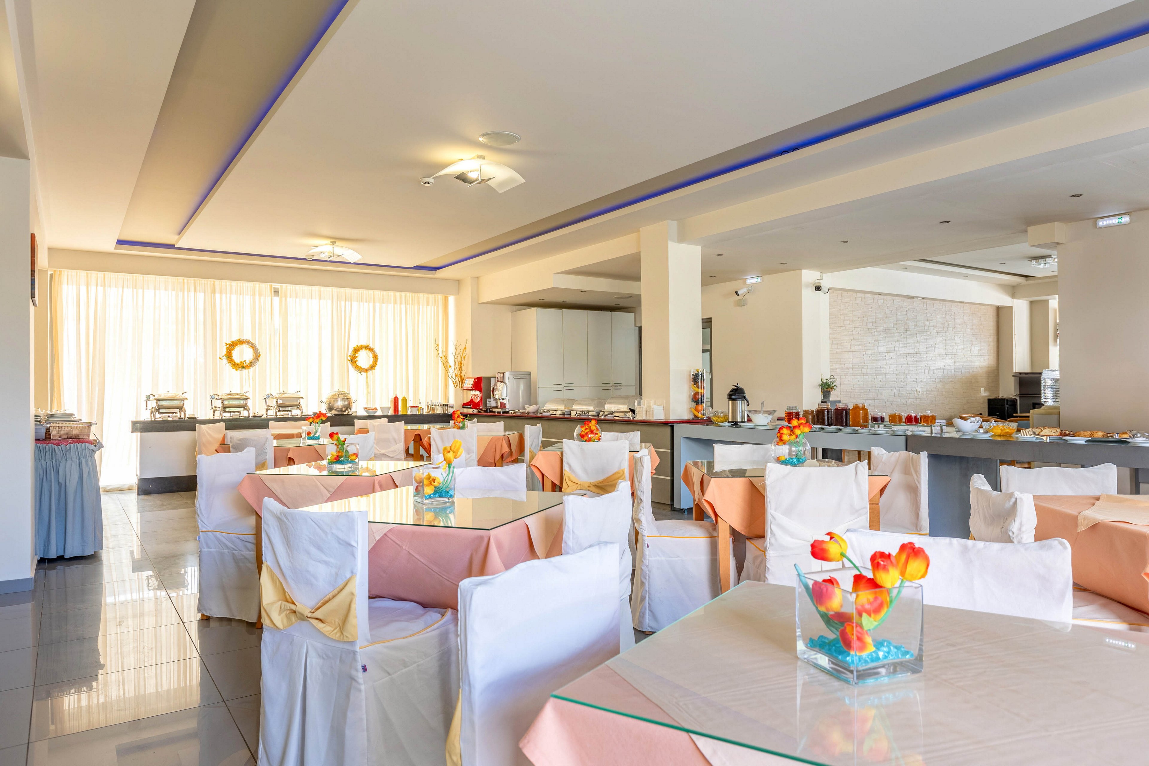 A modern hotel dining room is arranged with neatly covered tables and chairs, each decorated with pastel-colored cloths and bows. The space is brightly lit by natural light streaming through large sheer curtains at the back. A long buffet counter in the background is prepared with chafing dishes and various breakfast items. Glass vases with colorful artificial flowers add a cheerful accent to the otherwise neutral interior.