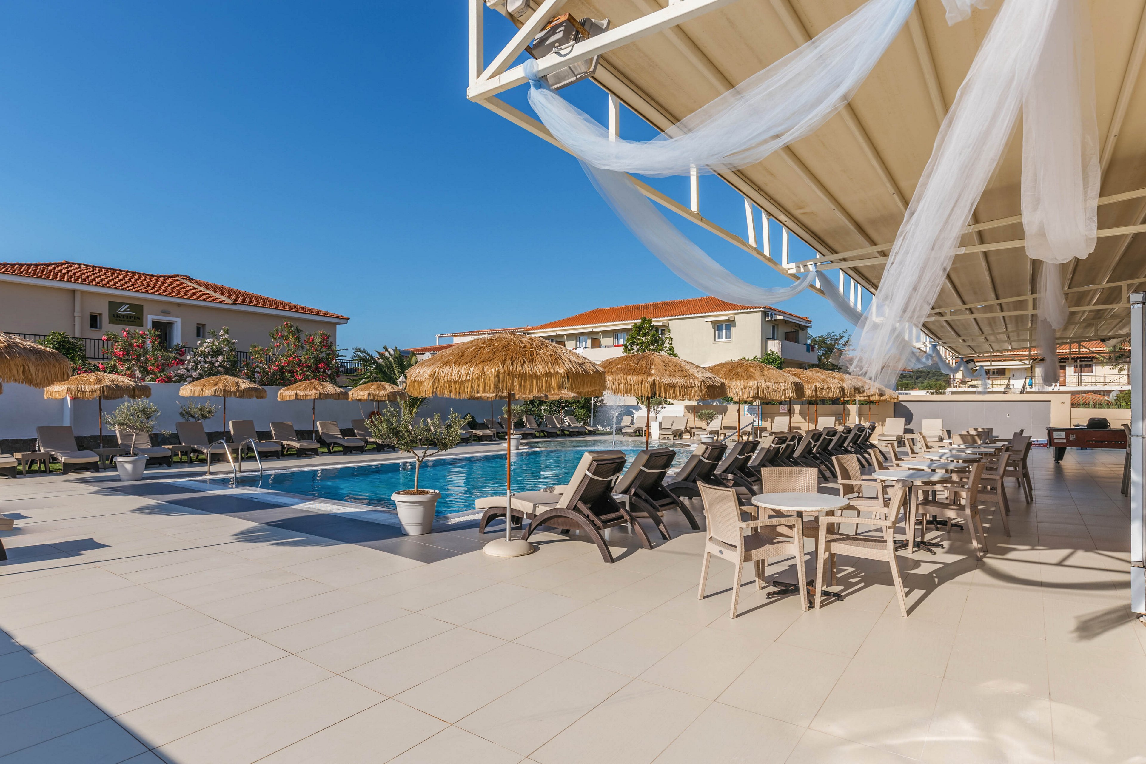 An outdoor resort pool area is shown under a clear blue sky. Numerous sun loungers line the pool, shaded by straw parasols and surrounded by potted plants. A covered terrace with flowing white fabric provides additional seating at tables and chairs. Low-rise buildings with red-tiled roofs frame the background, creating a relaxed vacation atmosphere.