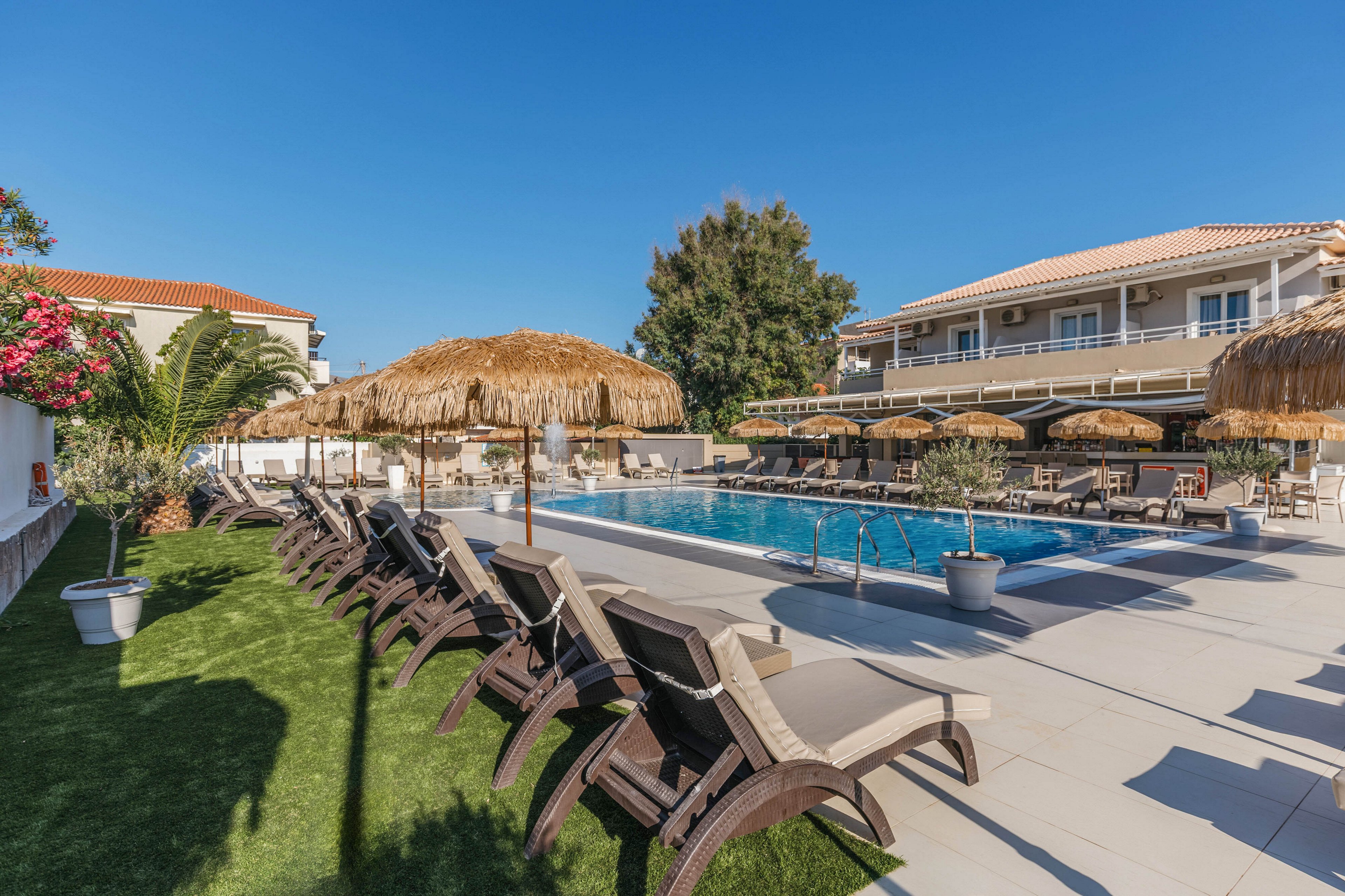 An outdoor resort swimming pool is surrounded by rows of reclining lounge chairs and straw parasols. The scene is set on a sunny day with clear blue skies and bright natural light. A mix of tiled deck and artificial grass areas frame the pool, with potted plants and flowering shrubs adding greenery. Two-story buildings with balconies overlook the pool, creating a relaxed vacation atmosphere.