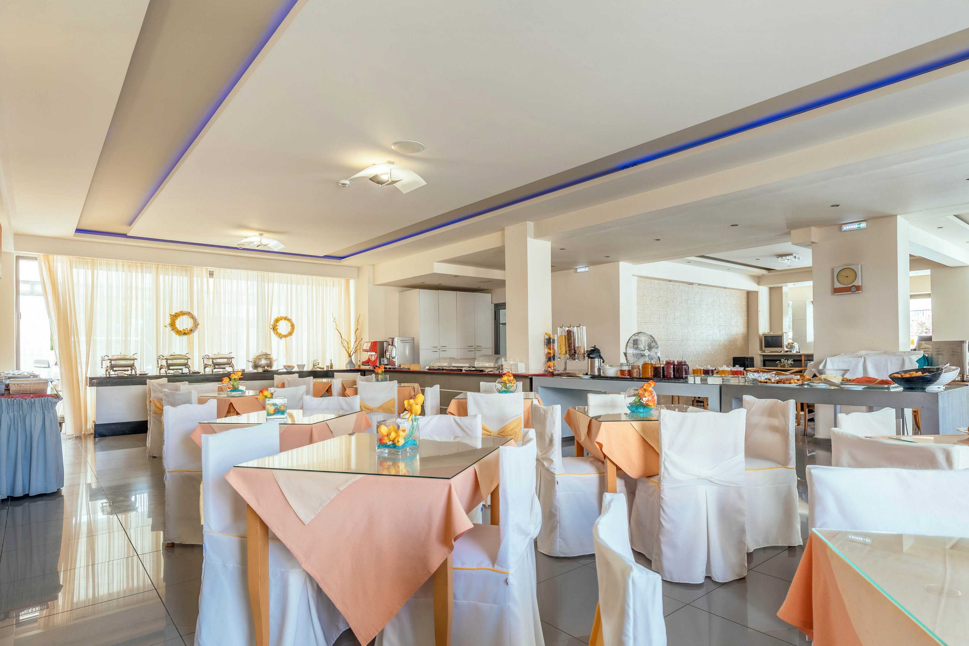 Spacious hotel restaurant interior with neatly arranged tables covered in white cloths and peach table runners. The room features a buffet counter in the background with various food and beverage stations. Large windows with sheer curtains allow natural light to fill the space, highlighting the glossy tiled floor. Decorative centerpieces and soft ceiling lighting create a clean, welcoming dining atmosphere.