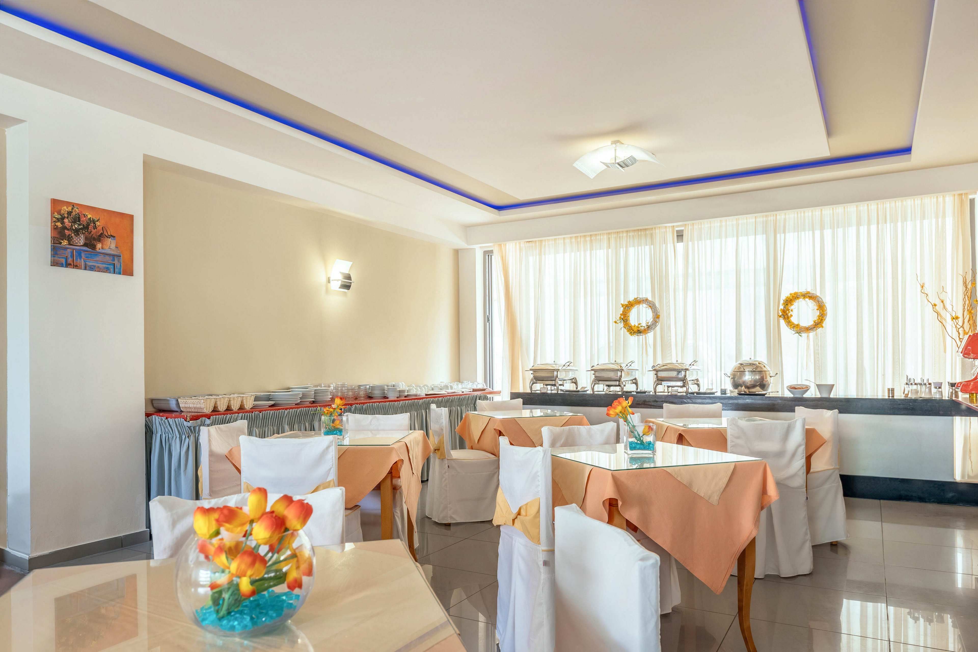 A modern hotel dining room is arranged with neatly covered tables and peach-colored tablecloths. White chair covers and soft neutral walls create a clean, elegant atmosphere. In the background, a buffet line with chafing dishes and serving trays is set up along a counter. Large windows with sheer curtains allow natural light to fill the space, highlighting decorative wreaths and a colorful floral centerpiece.