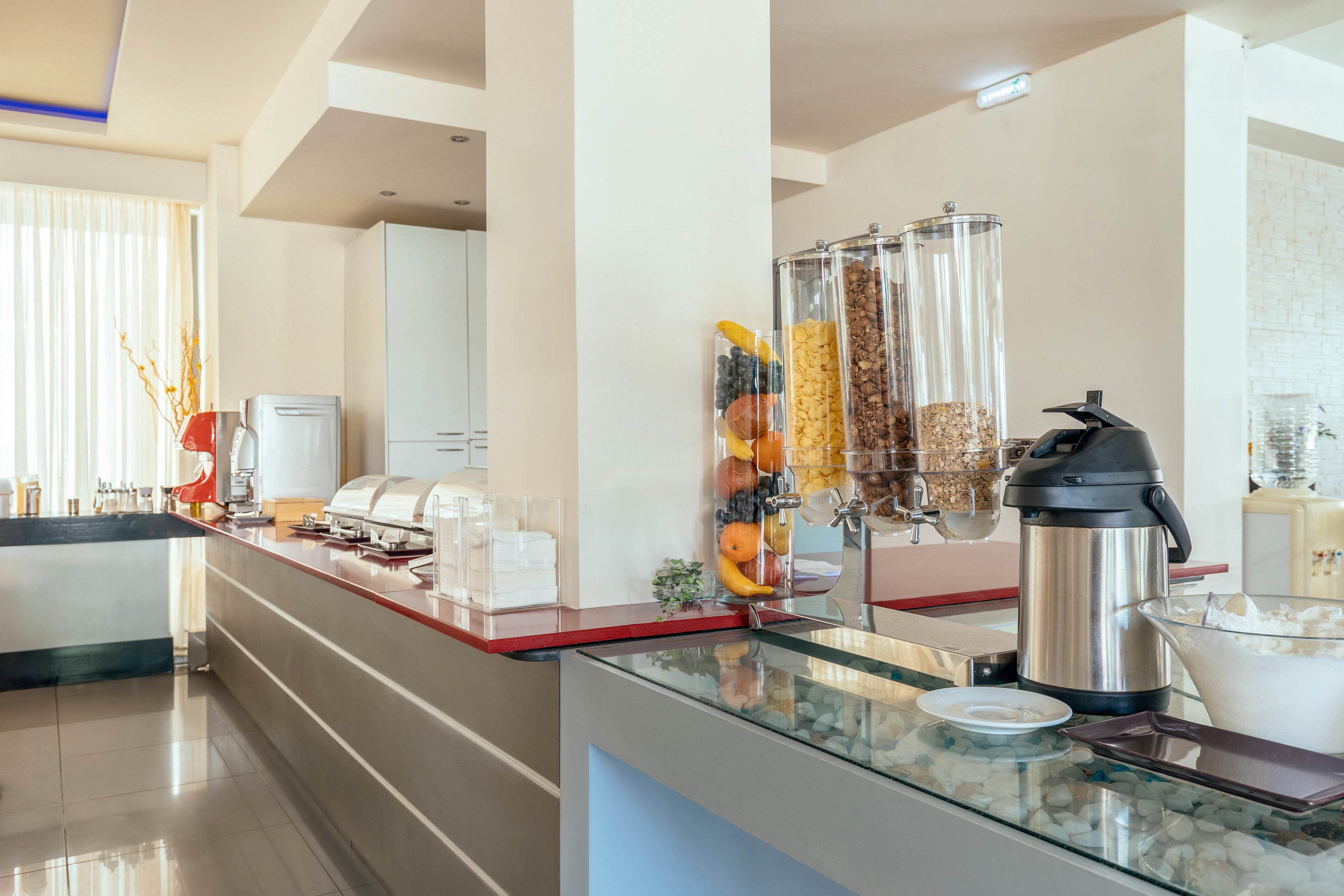 A contemporary self-service breakfast buffet is set up on a sleek counter in a bright dining area. Large transparent dispensers hold various cereals beside a tall container of fresh fruit. A stainless steel beverage dispenser and covered serving trays line the countertop, ready for guests. Soft natural light filters through sheer curtains, highlighting the clean, minimalist design of the space.