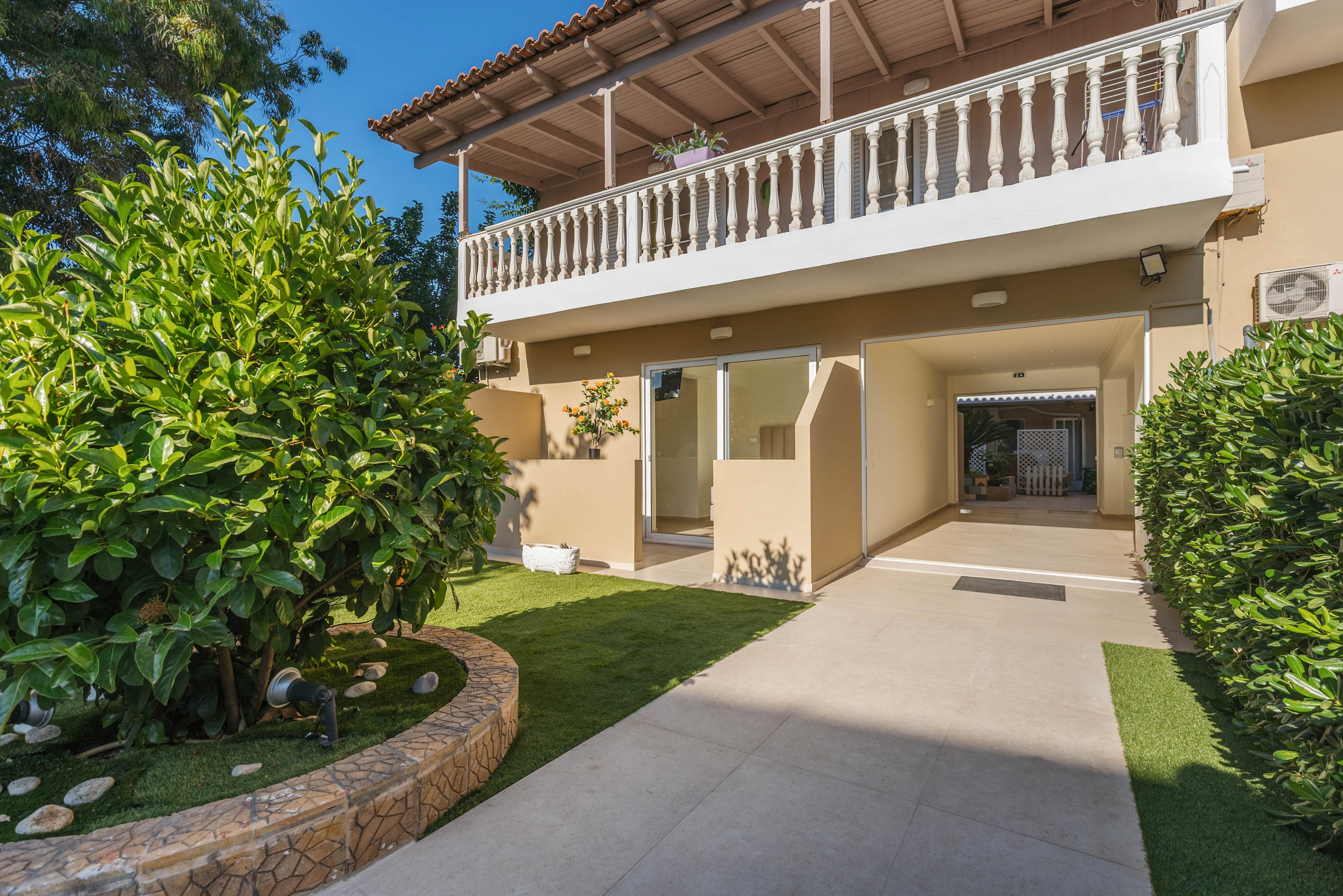 A bright residential courtyard leads to the entrance of a modern two-story house. The scene features a tiled walkway bordered by manicured green shrubs and a small circular stone garden bed. An upper balcony with white balustrades and a tiled roof overlooks the open ground-floor passageway. Warm beige walls and clear blue sky create a welcoming, sunlit atmosphere.