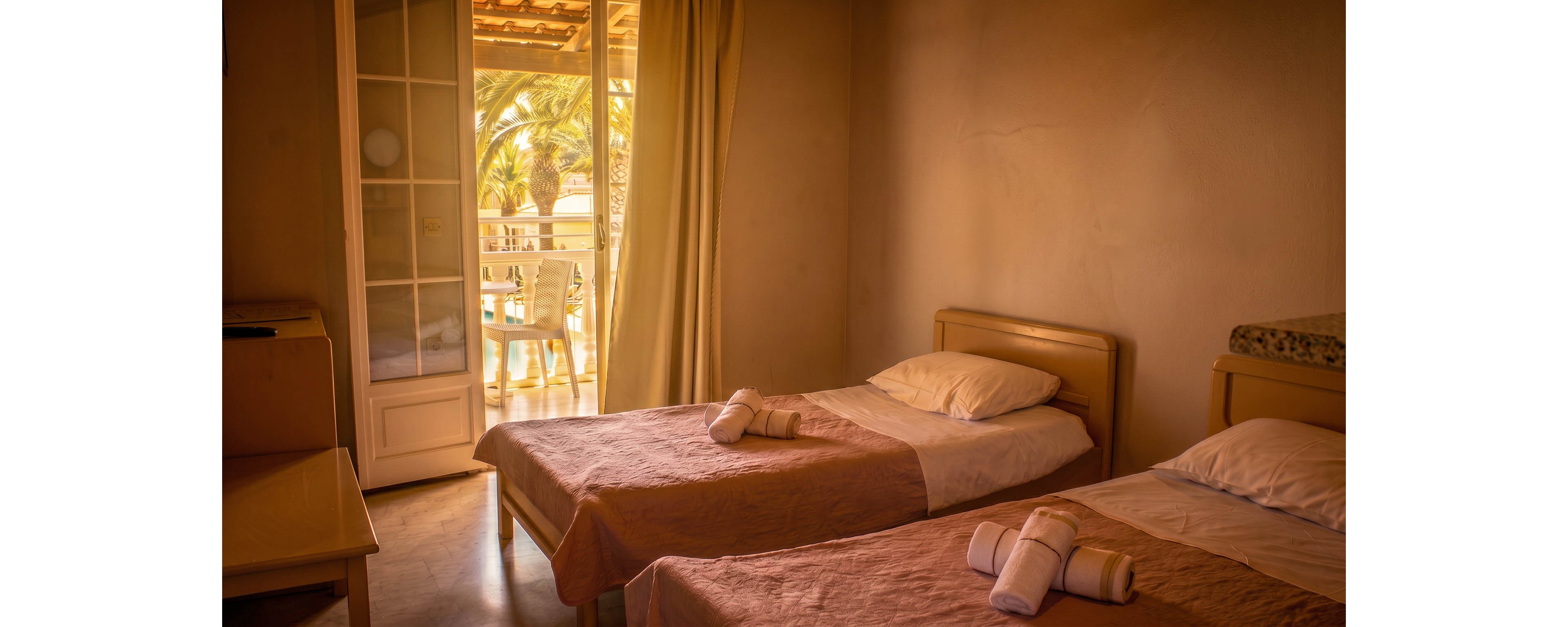A warm, softly lit room features two single beds neatly made with folded towels placed on top. The space opens onto a balcony through glass doors, allowing natural light to stream in. Outside, palm trees and a small seating area suggest a relaxing vacation setting. The overall atmosphere feels calm, inviting, and ideal for a peaceful stay.