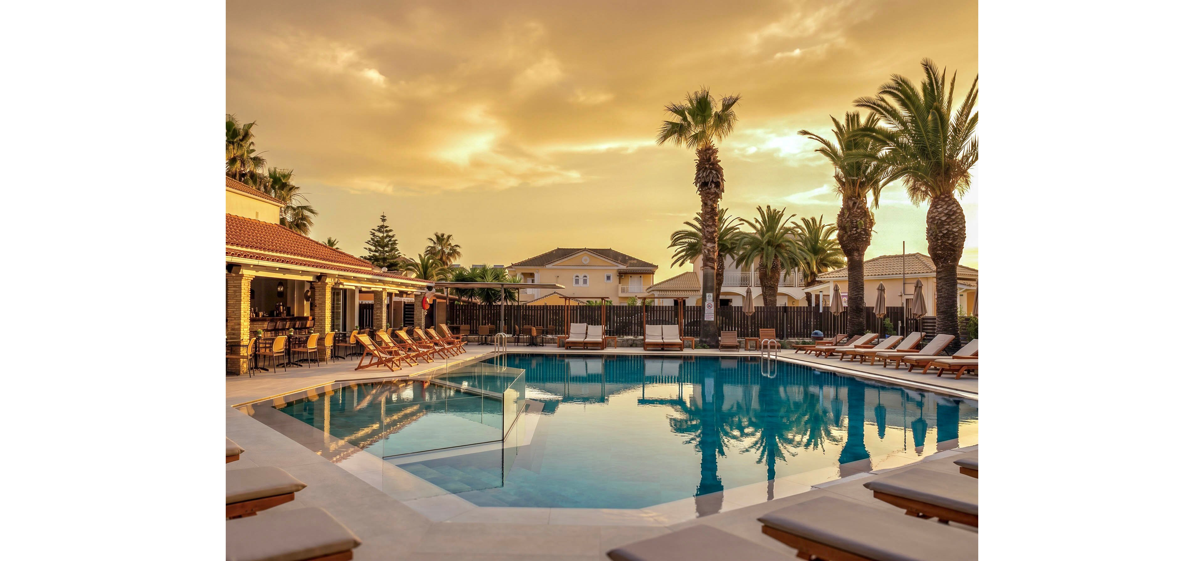 A tranquil resort swimming pool is shown at sunset, reflecting palm trees and warm golden clouds in the still water. Lounge chairs line the poolside, many arranged under umbrellas near a covered outdoor bar or seating area. Low residential-style buildings surround the pool, giving the scene a private, exclusive atmosphere. The overall mood is calm and inviting, suggesting a relaxing vacation or evening retreat.