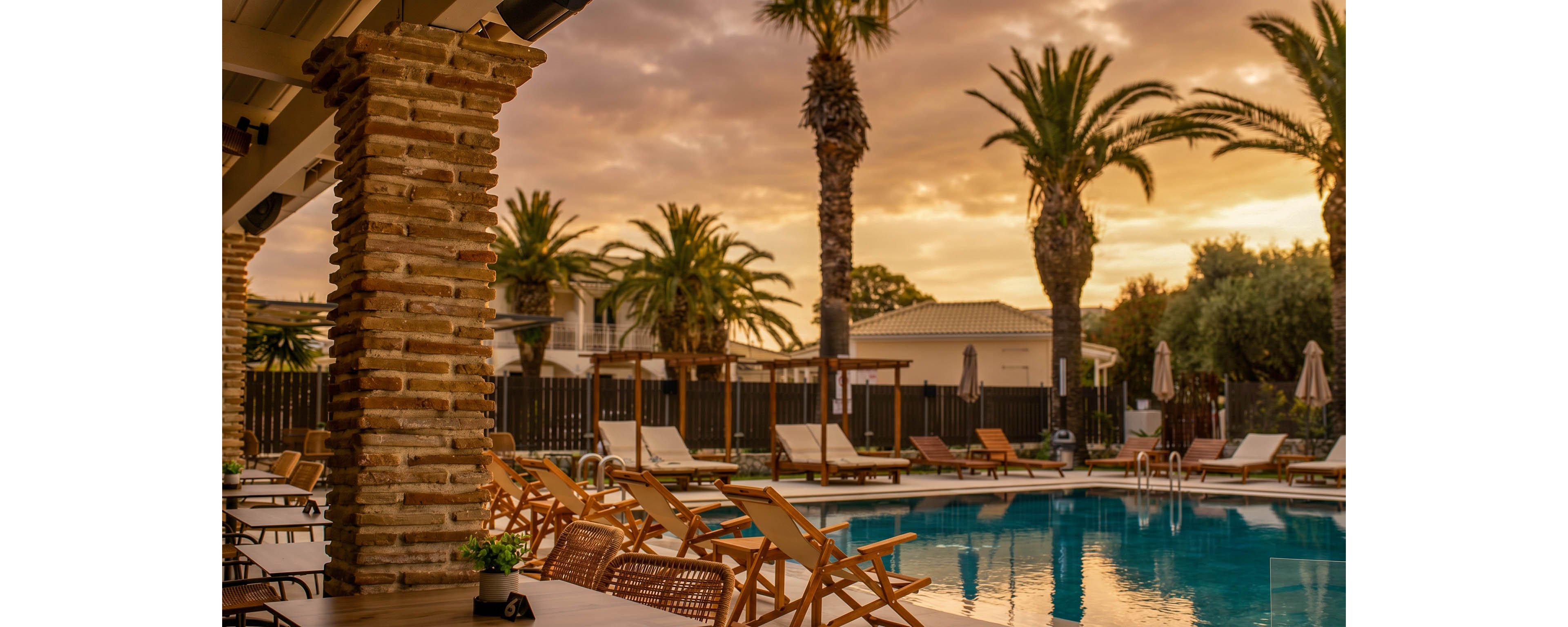 A tranquil resort swimming pool is surrounded by wooden lounge chairs and umbrellas at dusk. Tall palm trees frame the scene, reflecting softly in the calm blue water. Warm golden light from the sunset casts a glow over the pool deck and nearby buildings. The empty seating and serene atmosphere suggest a relaxing vacation setting.