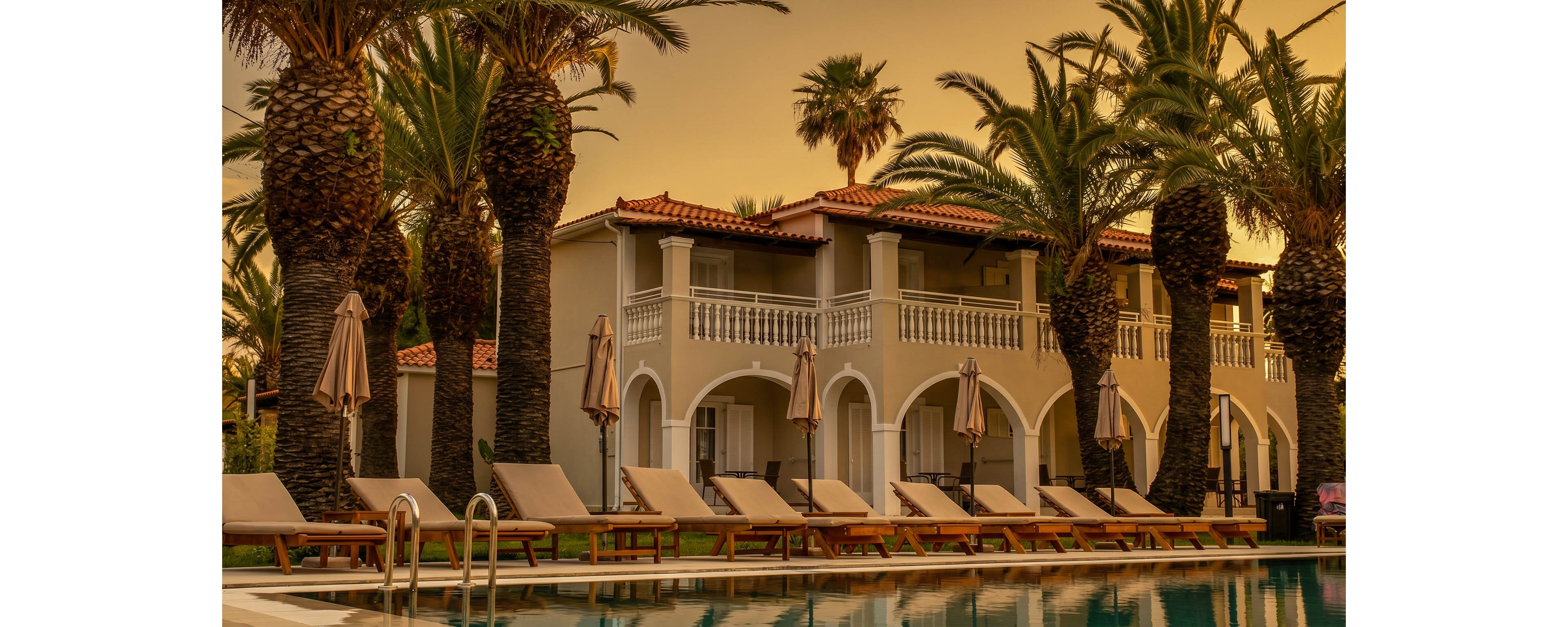 A Mediterranean-style resort building stands behind a tranquil swimming pool at sunset. The structure features arched ground-floor terraces, upper balconies, and a red-tiled roof. Tall palm trees line the poolside, casting warm shadows across the loungers and closed umbrellas. The golden sky and reflections in the water create a calm, luxurious vacation atmosphere.