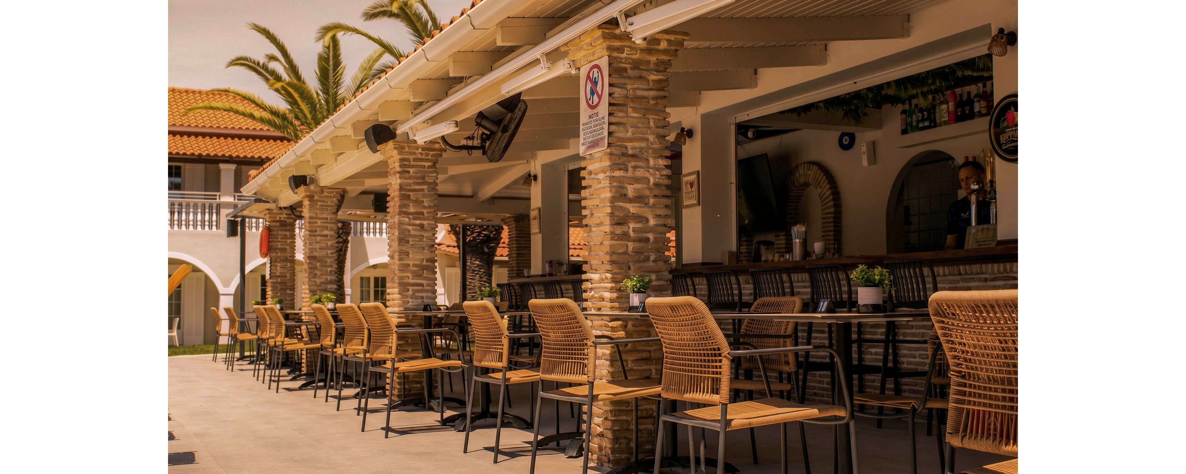 An open-air cafe features rows of wicker chairs and high tables under a covered patio. Brick columns support the roof, creating a shaded walkway beside the seating area. Potted plants and bar counters add a casual, welcoming atmosphere. Warm sunlight and palm trees in the background suggest a Mediterranean or coastal resort setting.
