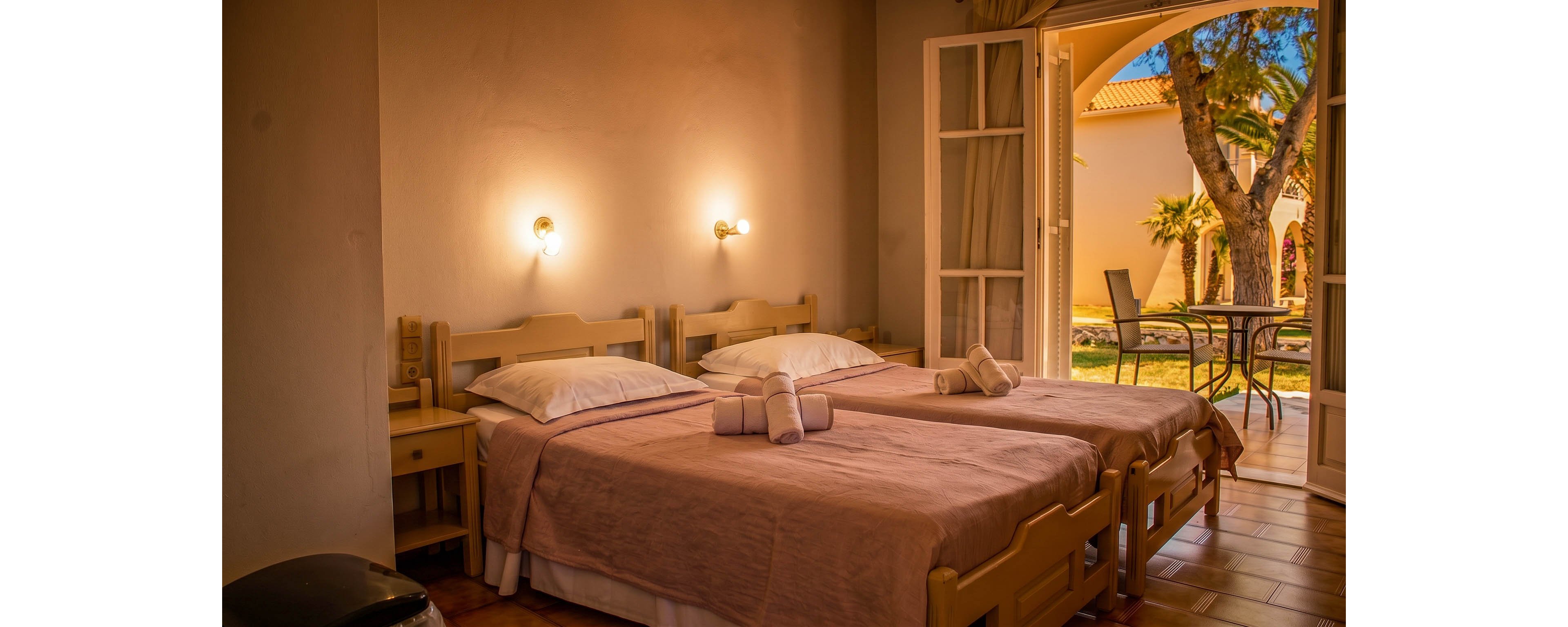 A warm, inviting bedroom features two twin beds neatly made with soft pink bedding and folded towels placed on top. Soft wall lights cast a gentle glow over wooden furniture, including bedside tables and a small desk. French doors open to a sunlit patio with a round table, chairs, and views of trees and a building with a tiled roof. The overall scene suggests a comfortable guest room or vacation accommodation with indoor-outdoor living.