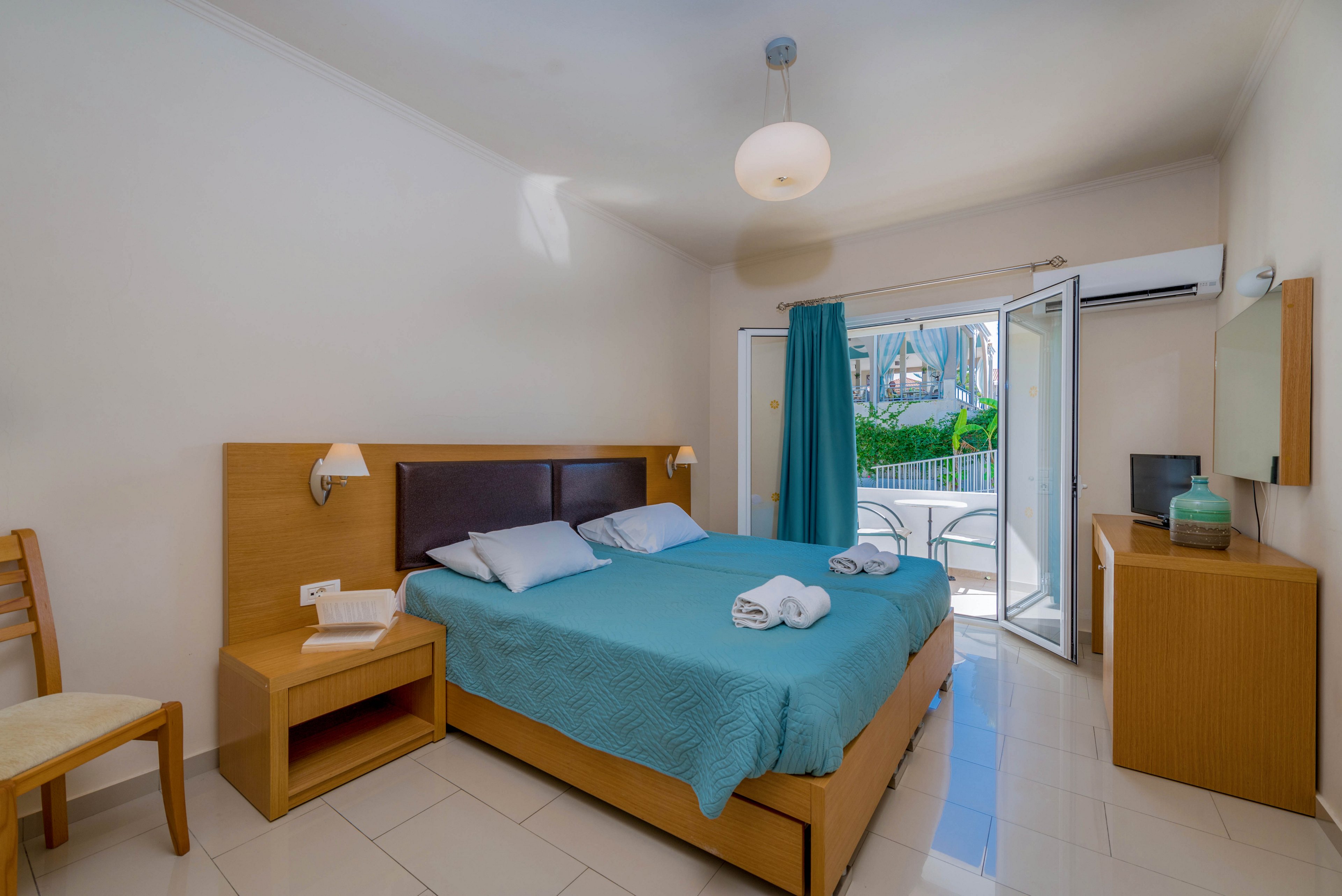 A tidy hotel-style bedroom features a double bed with a turquoise quilt and neatly folded white towels. Light wood furniture, including bedside tables, a chair, and a small desk with a mirror, complements the neutral walls and tiled floor. Natural light streams in through open glass doors leading to a balcony with greenery outside. A ceiling light, wall lamps, and an air conditioner above the door complete the comfortable, modern interior.