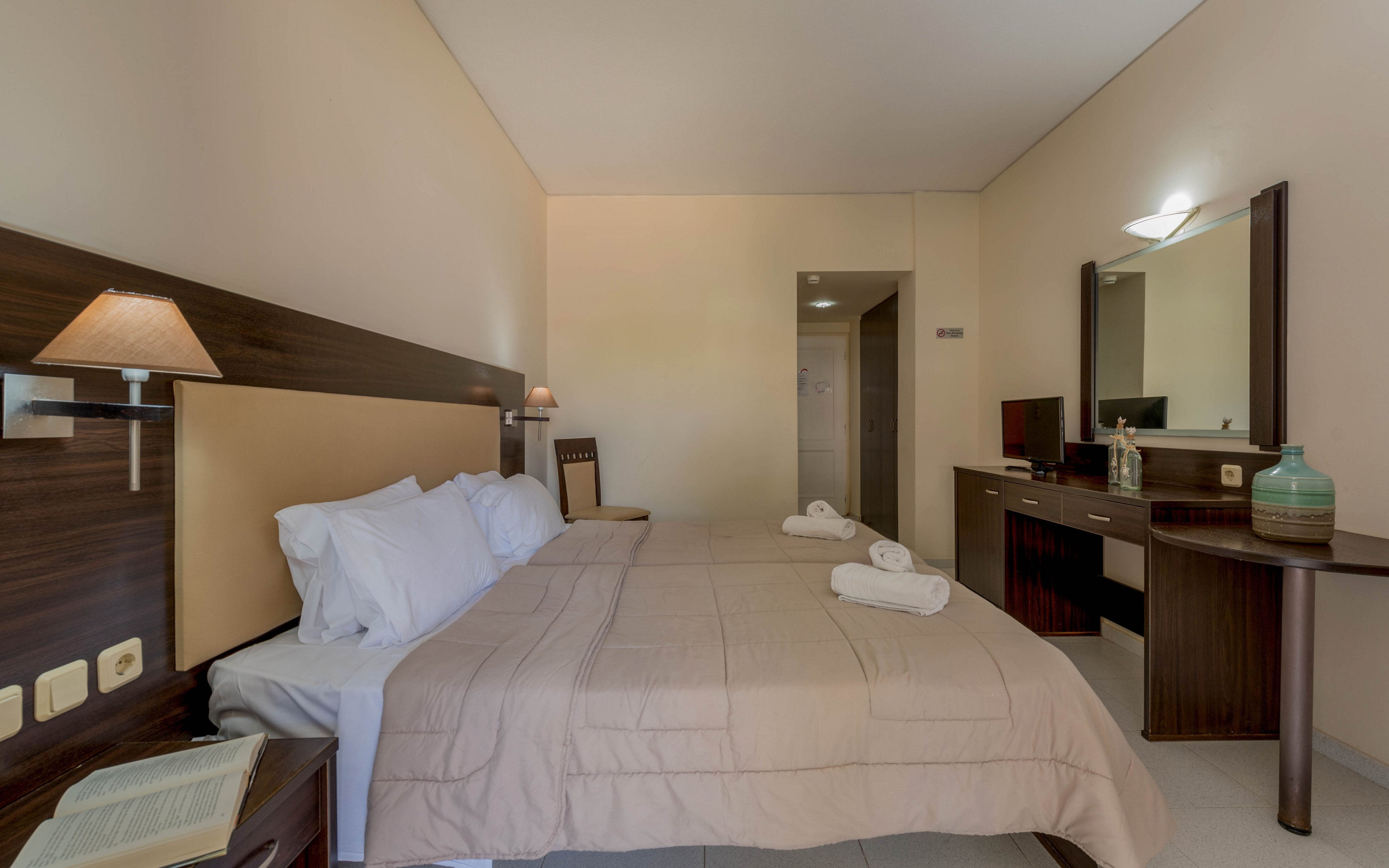 A tidy hotel bedroom features a double bed with beige bedding and white pillows. Rolled white towels are placed neatly on the bed, and a book lies open on the bedside table. The room includes a wooden desk with a mirror, chair, and decorative items, all in a neutral color palette. Soft wall lighting and clean walls create a calm, minimalist atmosphere.
