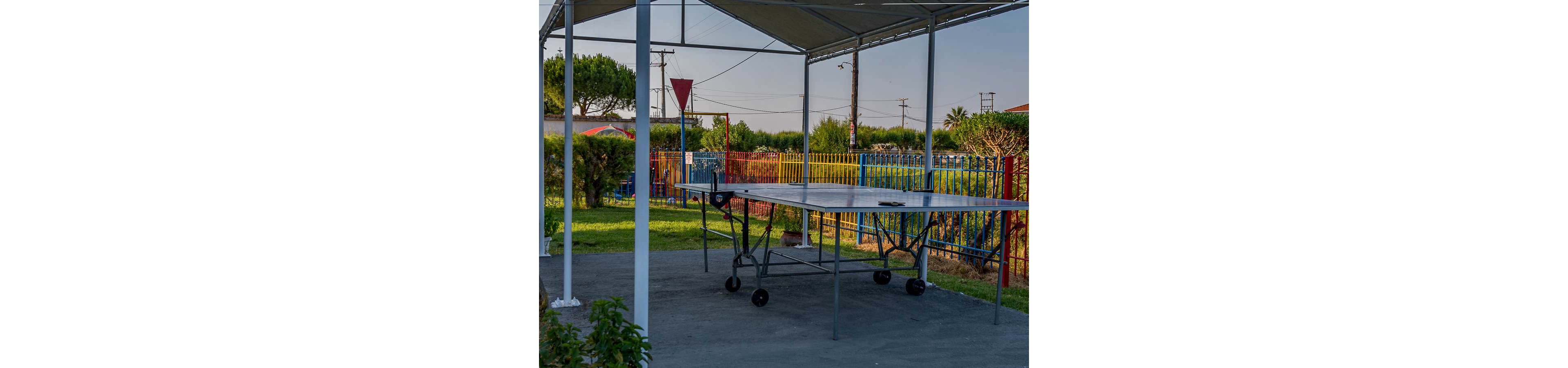 An outdoor ping pong table stands beneath a metal canopy on a paved surface. The scene is set in a fenced yard with colorful railings and green bushes. Utility poles and wires are visible in the background, suggesting a suburban or residential area. The lighting indicates a calm, late afternoon or early evening atmosphere.