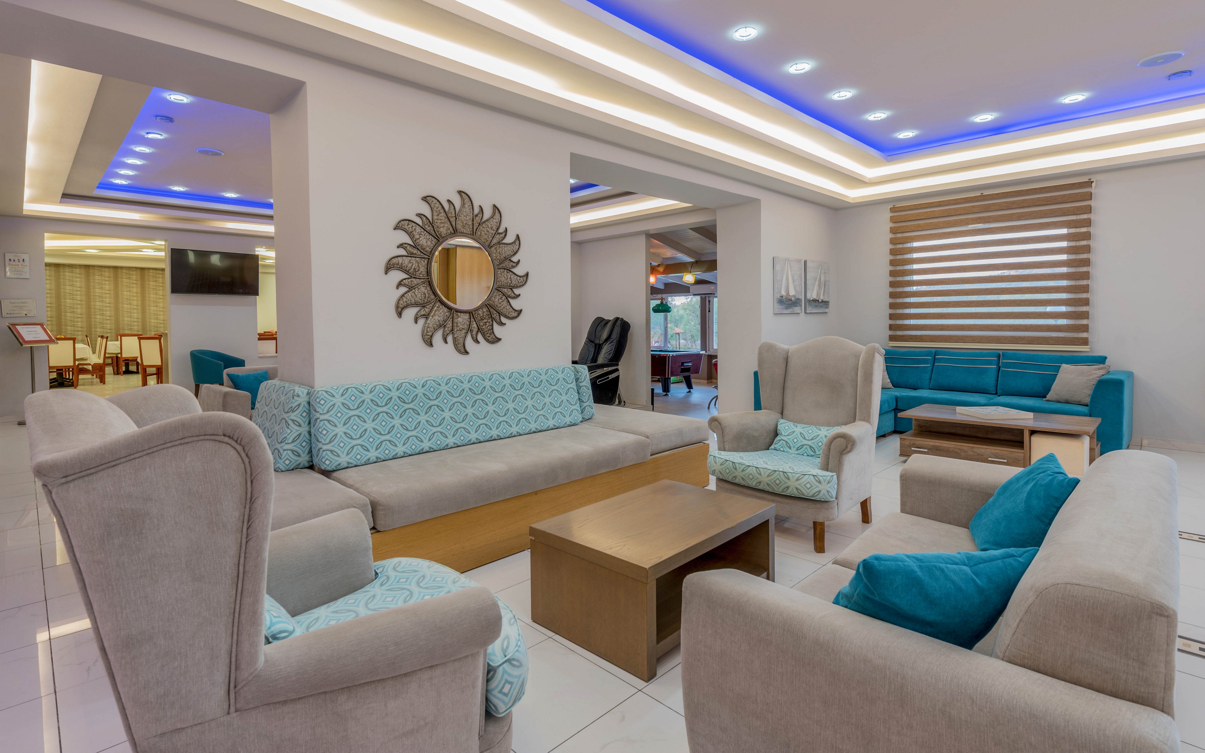 A contemporary lounge area features beige armchairs and sofas accented with turquoise cushions and upholstery. The space is brightly lit with recessed ceiling lights and blue LED strips, creating a clean, modern atmosphere. A decorative sunburst wall mirror and abstract artwork add visual interest, while large windows and blinds frame the seating area. The room appears organized and inviting, suitable for hotel guests or visitors to relax and socialize.