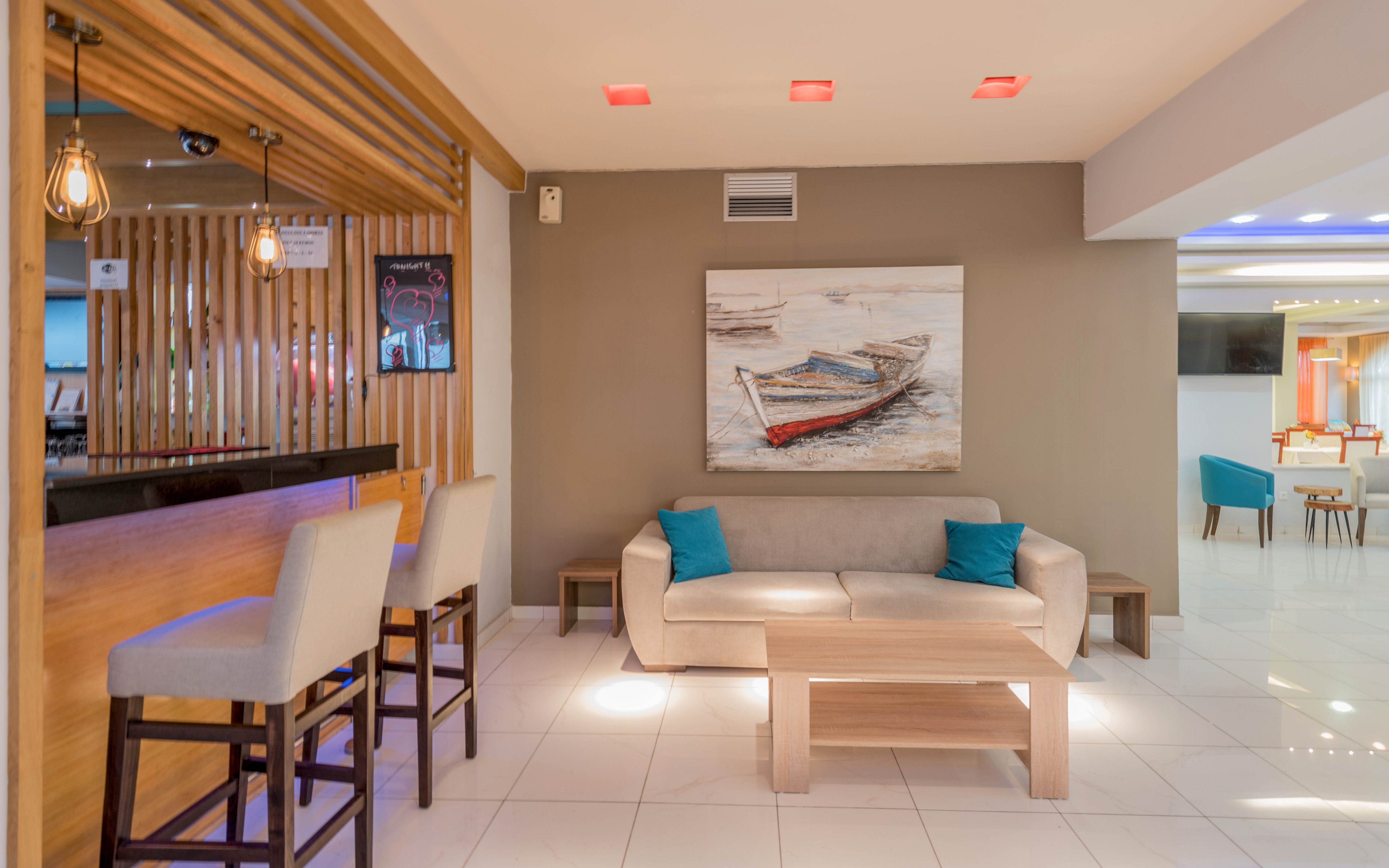 Contemporary lounge area featuring a beige sofa with turquoise accent pillows and a light wood coffee table. A large wall painting of a boat on the water serves as the focal point against a neutral-colored wall. To the left, a sleek bar with wooden slats and pendant lights offers high stools for seating. In the background, additional chairs and tables create a bright, open social space with clean white flooring.
