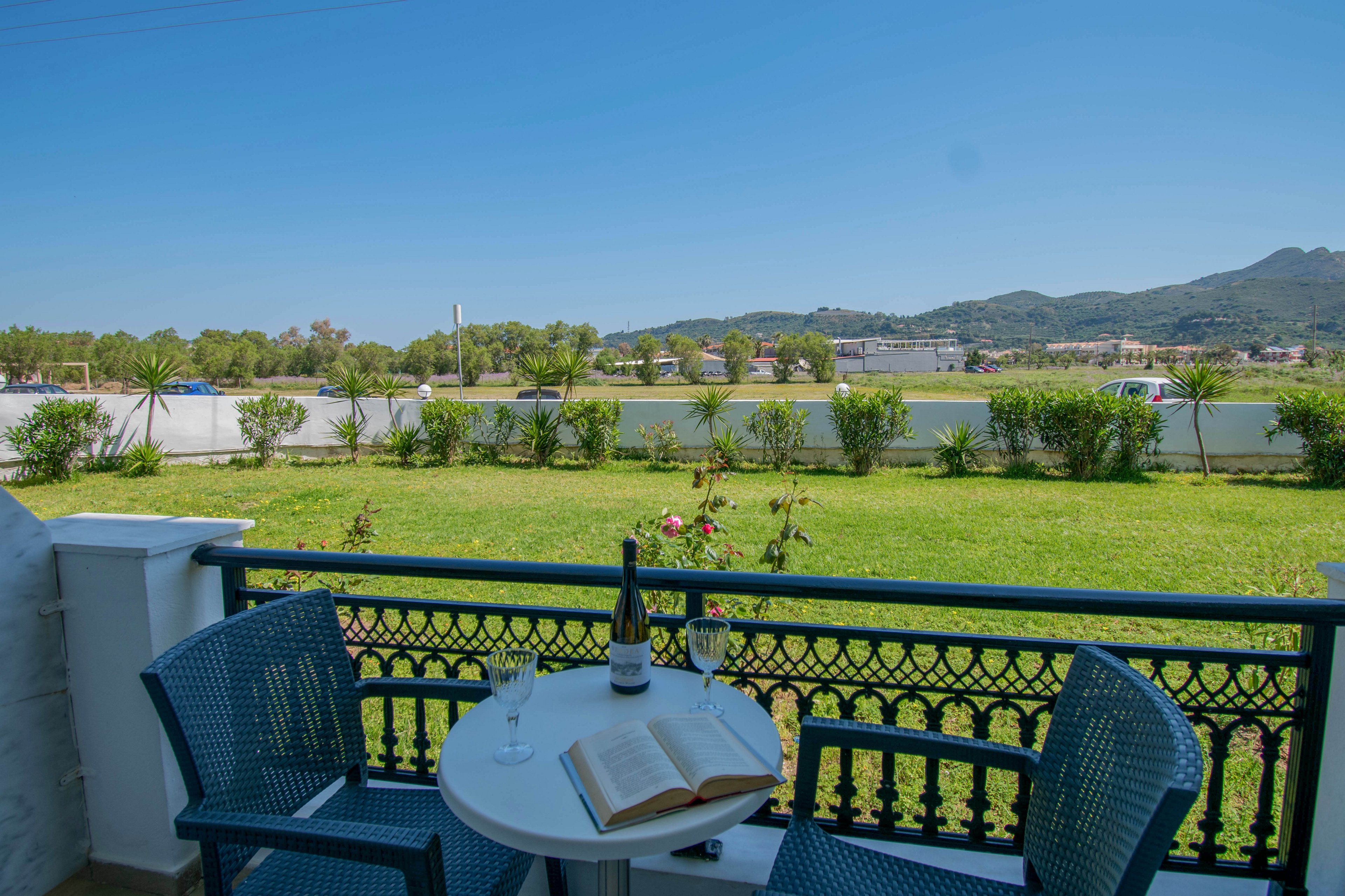A small balcony overlooks a green lawn and distant hills under a clear blue sky. On the round white table sit a wine bottle, a filled wine glass, and an open book, suggesting a quiet moment of relaxation. Two blue chairs frame the table, and decorative railing details add character to the scene. In the background, trees, buildings, and mountains complete the peaceful outdoor setting.