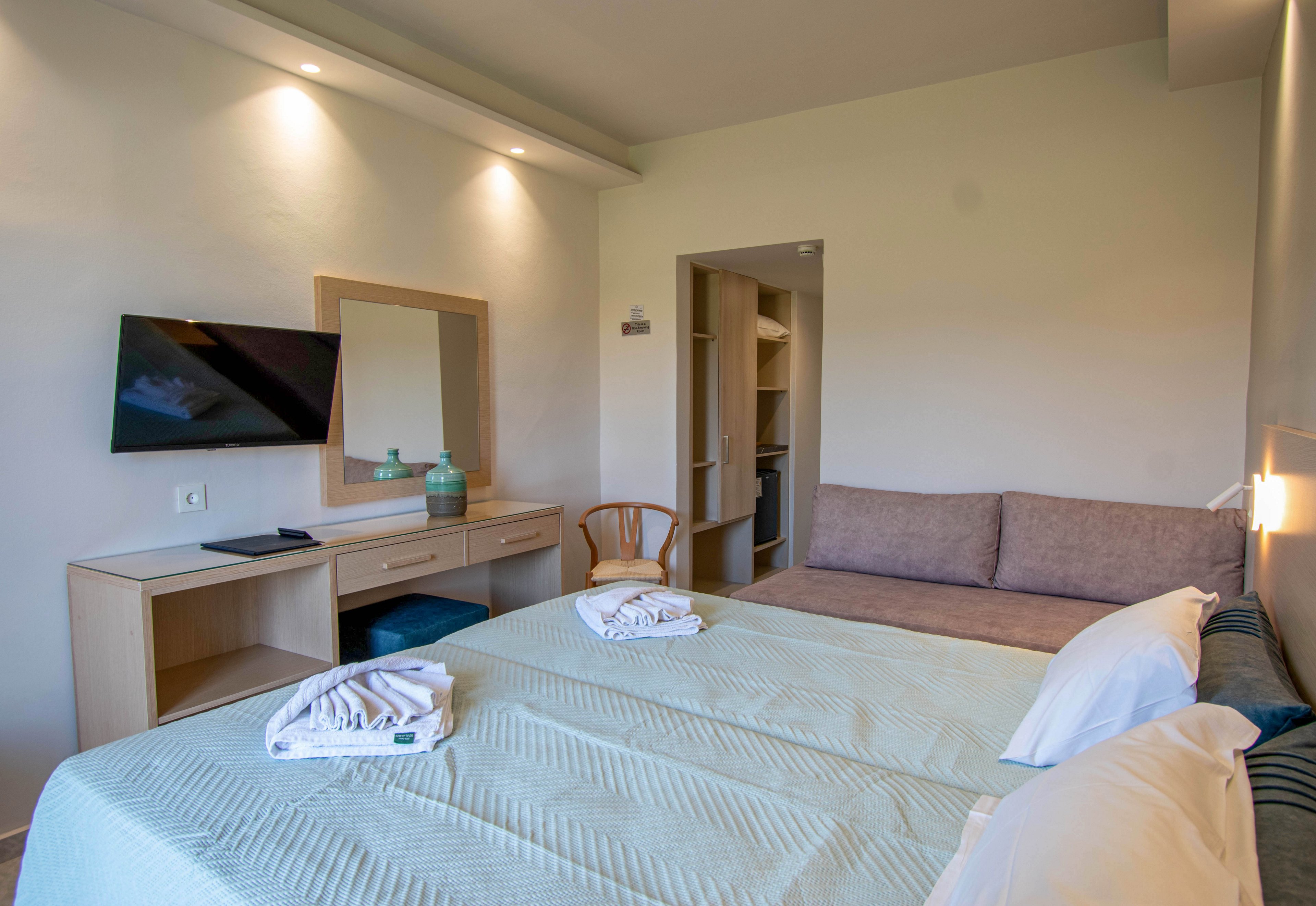 A contemporary hotel room features two neatly made twin beds with folded white towels placed on top. A wall-mounted flat-screen television faces a light wood desk and mirror, with decorative vases adding a touch of color. A cushioned sofa sits against the wall near an open doorway leading to a small hallway or closet area. Soft, neutral tones and recessed ceiling lights create a calm, inviting atmosphere suitable for guests.