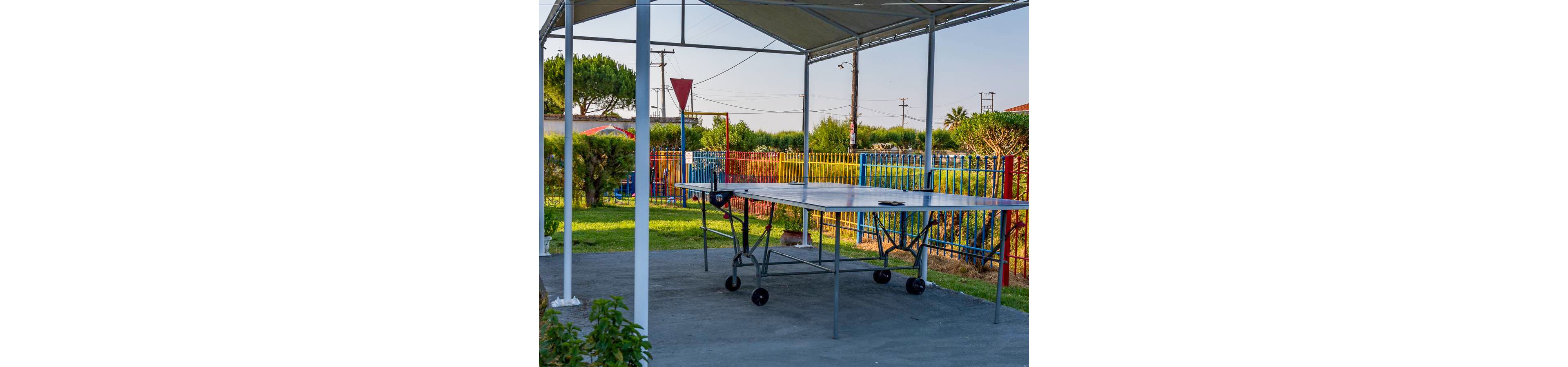 An outdoor ping pong table stands under a metal canopy in a small park or recreational area. Colorful fencing and playground structures are visible in the background, along with trees and utility poles. The scene is lit by warm daylight, giving a relaxed neighborhood atmosphere. The setting suggests a casual space for games and social activities.