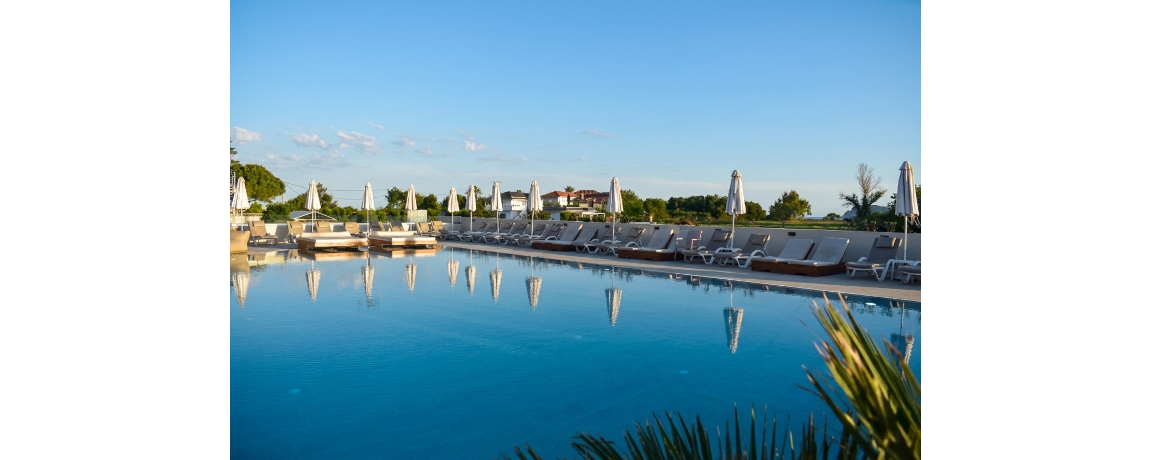 A large outdoor swimming pool reflects the clear blue sky at a resort. Numerous sun loungers and closed umbrellas line the poolside, suggesting a quiet, early or late day setting. Buildings and greenery appear in the background, adding depth to the scene. The calm water and soft light create a tranquil, vacation-like atmosphere.