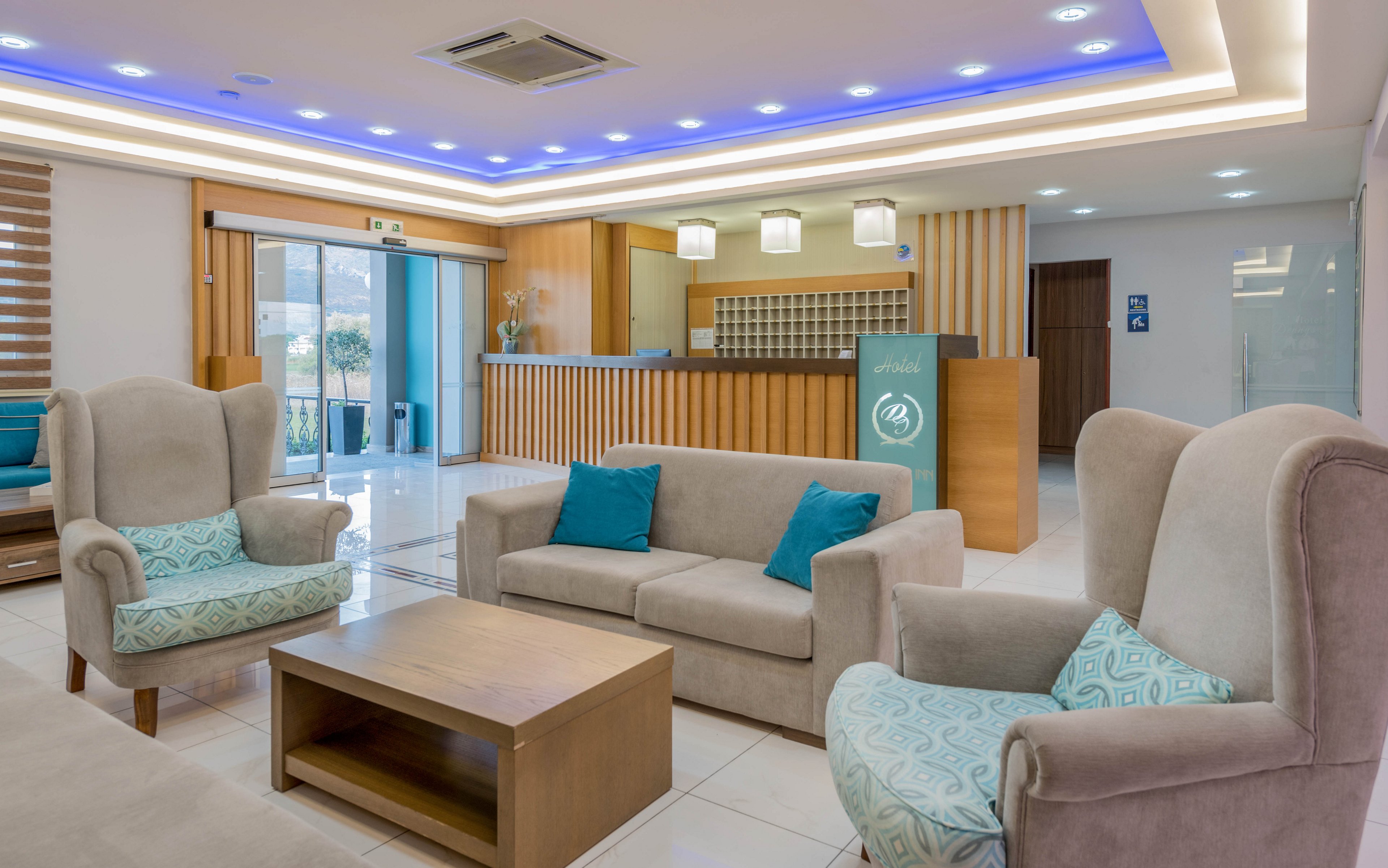 A contemporary hotel lobby features beige armchairs and a sofa arranged around a wooden coffee table. The reception desk stands in the background with pendant lights above and decorative wood paneling. Blue accent pillows and patterned cushions add color to the neutral interior. Recessed ceiling lighting and large windows create a bright, welcoming atmosphere.