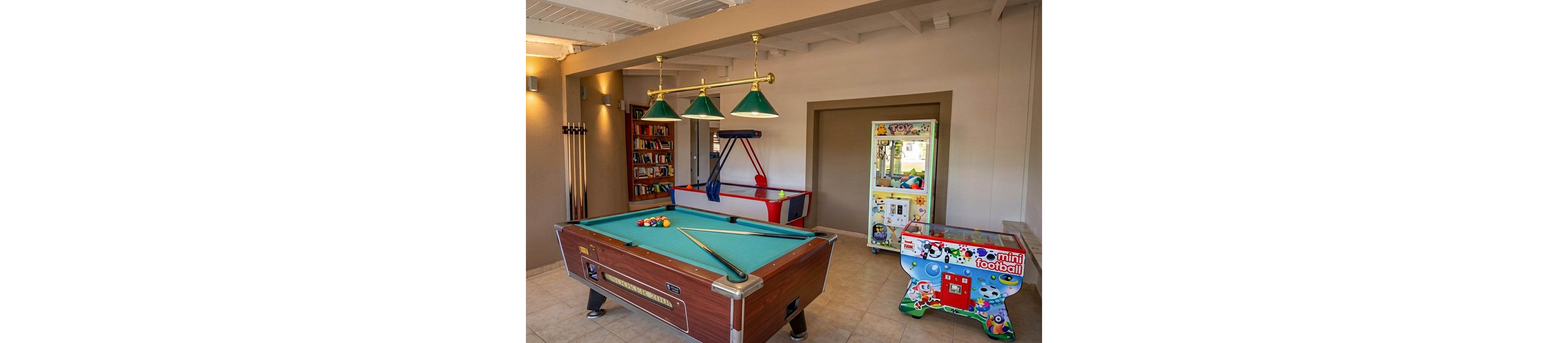 A recreational game room features a central pool table with cues and colorful balls arranged on its surface. Behind it, a bookshelf and wall-mounted cue rack add to the leisure setting. In the background are an air hockey table and a claw machine labeled "TOY" filled with plush prizes. A brightly decorated mini football arcade machine stands to the right, creating a fun, family-friendly atmosphere.