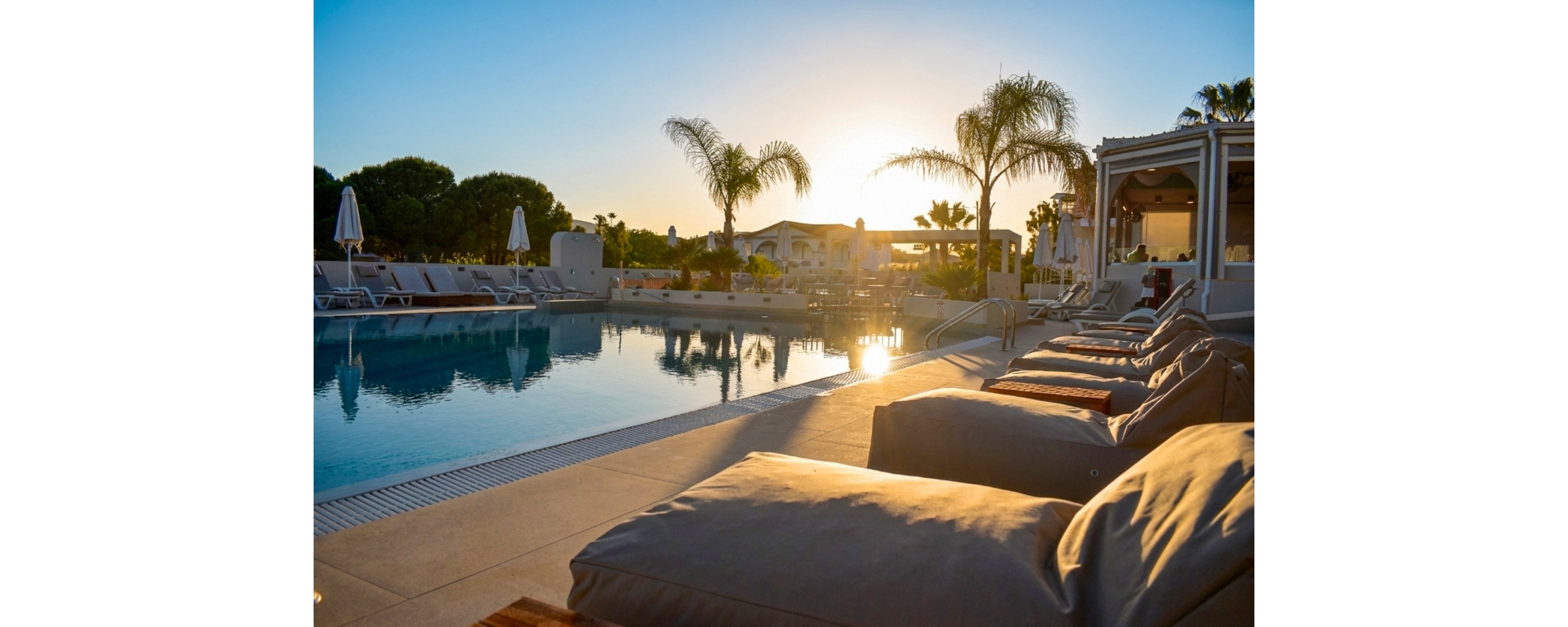 A row of cushioned loungers lines the edge of a tranquil outdoor swimming pool at a resort. The scene is captured at sunset, with warm golden light reflecting on the water’s surface. Palm trees and shaded seating areas frame the pool, creating a relaxed vacation atmosphere. The overall mood is serene and inviting, suggesting leisure and comfort in a modern holiday setting.