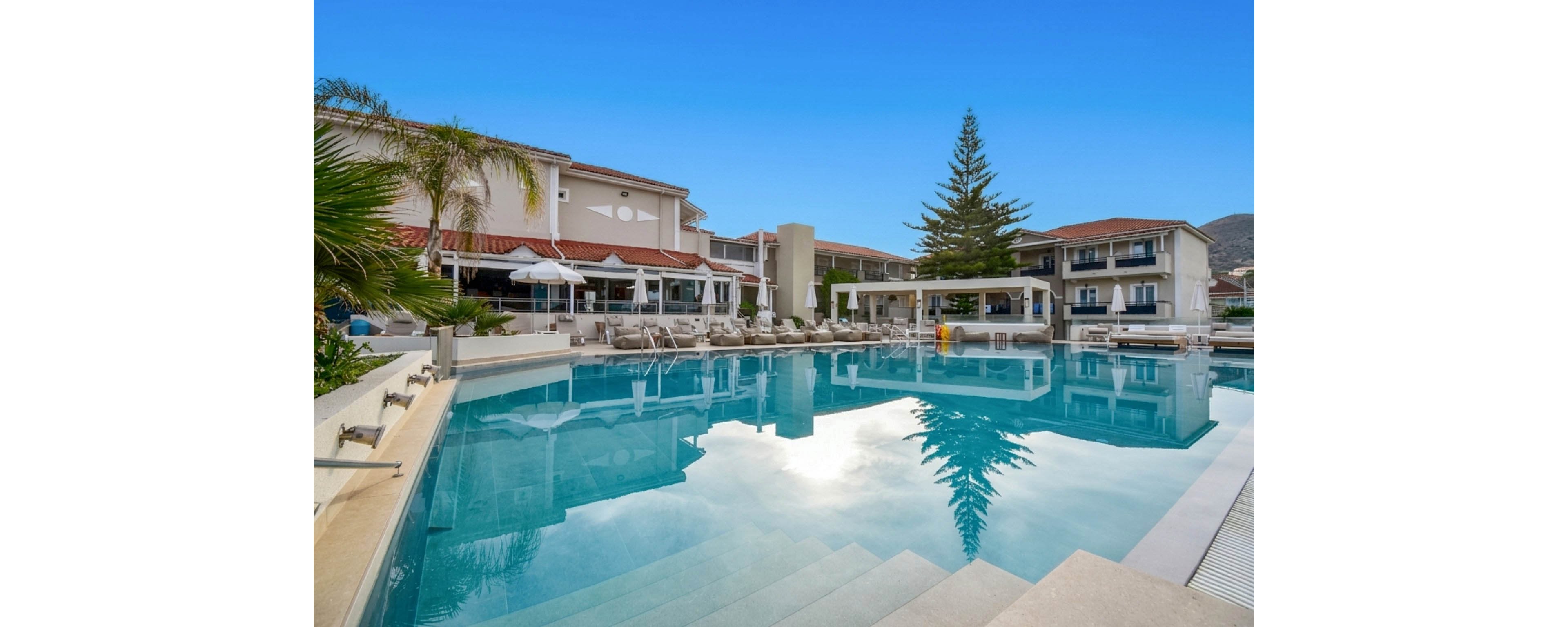 A large outdoor swimming pool reflects the surrounding resort buildings and bright blue sky. Sun loungers and umbrellas line the poolside terrace, suggesting a relaxing vacation setting. Palm trees and a tall evergreen add greenery around the water, while distant hills frame the background. The calm, clear water and modern architecture create a serene, upscale atmosphere.