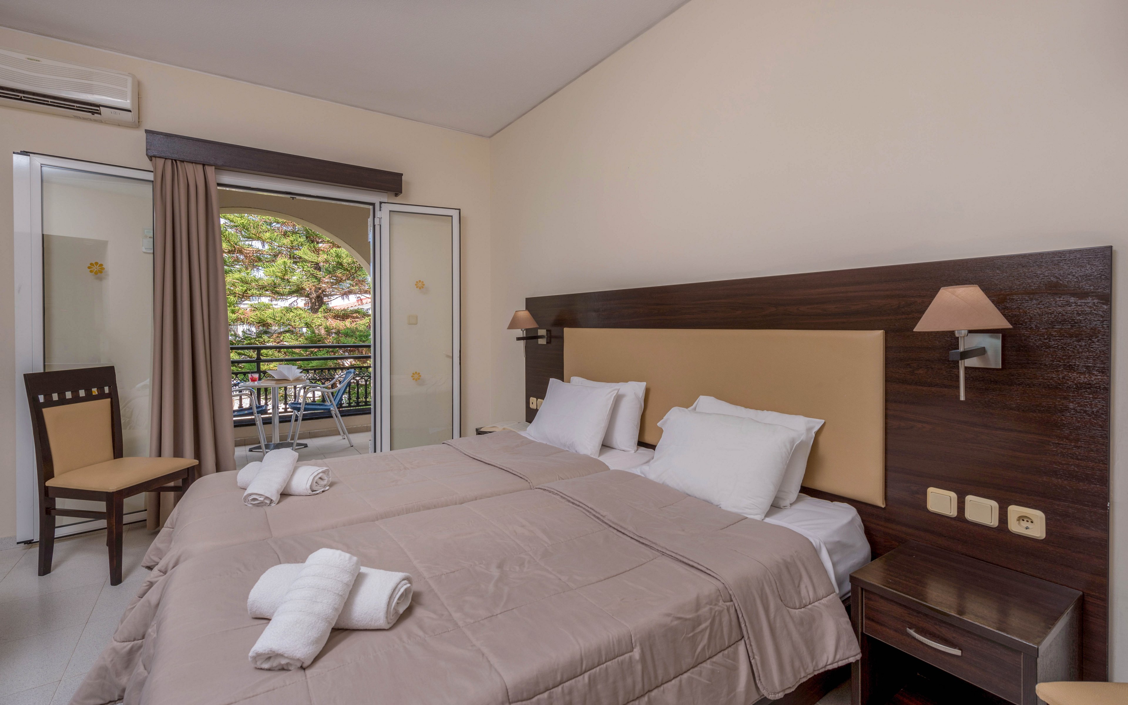 A neatly made hotel room features a large double bed with beige bedding and white pillows. Rolled white towels are placed on the bed, suggesting readiness for new guests. A sliding glass door opens to a balcony with a view of green trees outside. The room includes a wooden chair, bedside table, wall-mounted lamps, and a neutral color palette that creates a calm, inviting atmosphere.