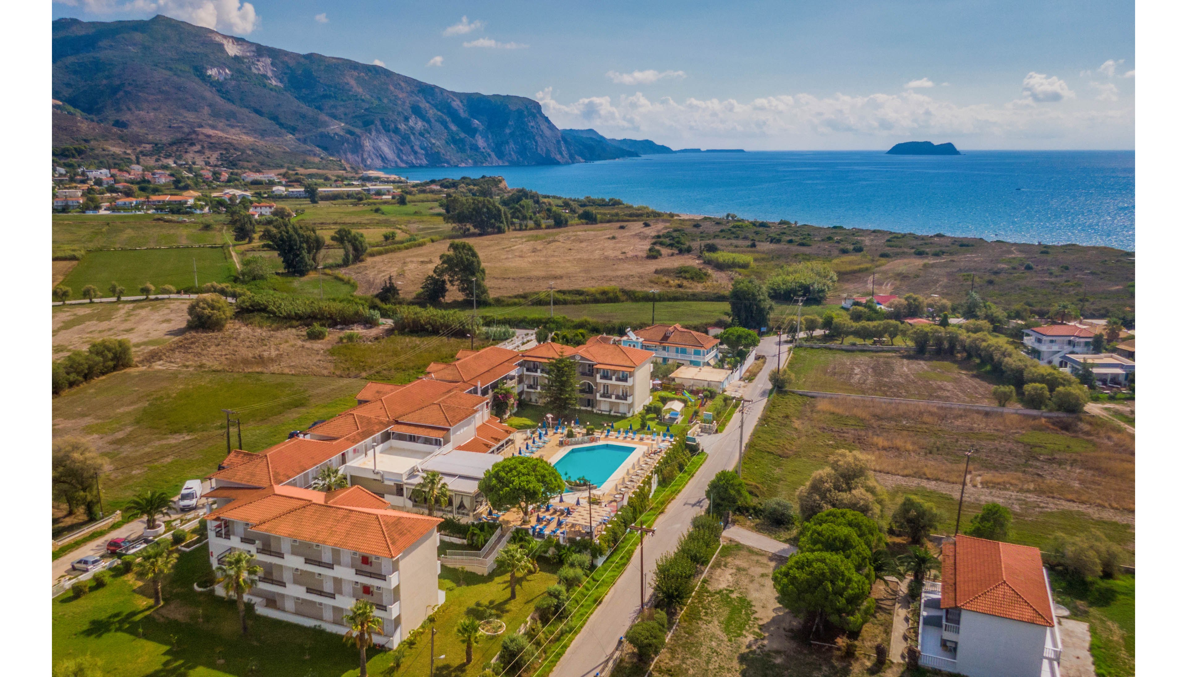 Aerial view of a coastal resort complex with red-roofed buildings and a central swimming pool. The property is surrounded by open fields and greenery, with a road running alongside it. In the distance, mountains rise along the shoreline, meeting the blue sea. The scene suggests a peaceful vacation destination with sunny weather and clear skies.