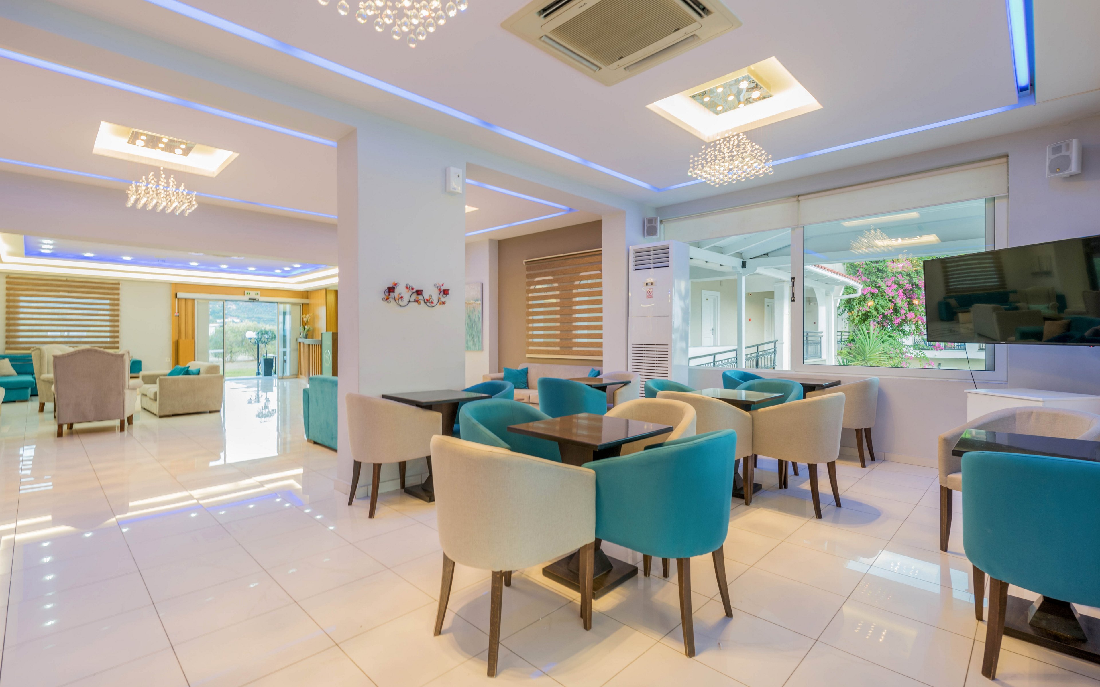 A bright, modern lounge area features turquoise and beige armchairs arranged around dark wooden tables. Large windows and glass doors let in natural light and offer views of an outdoor terrace with greenery. Recessed ceiling lights and contemporary chandeliers create a warm, inviting atmosphere. The clean white tile floor and neutral walls emphasize the colorful seating and sleek interior design.
