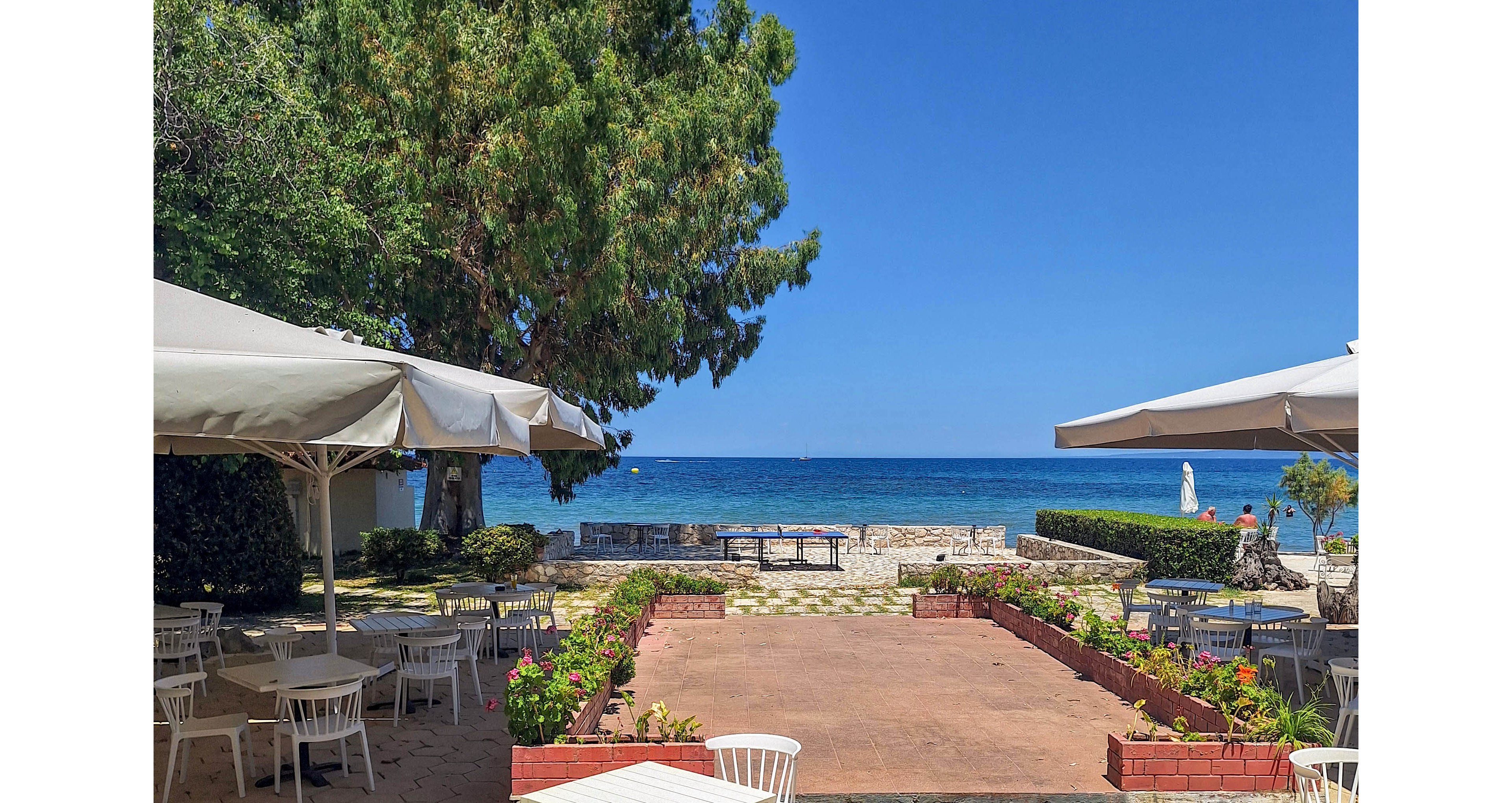 An outdoor café terrace overlooks a calm blue sea under a clear sky. White tables and chairs are arranged on a hexagonal-tiled patio, partially shaded by large umbrellas. A central walkway bordered by low flower beds leads toward the waterfront seating area. Lush green trees and shrubs frame the scene, creating a relaxed coastal atmosphere.