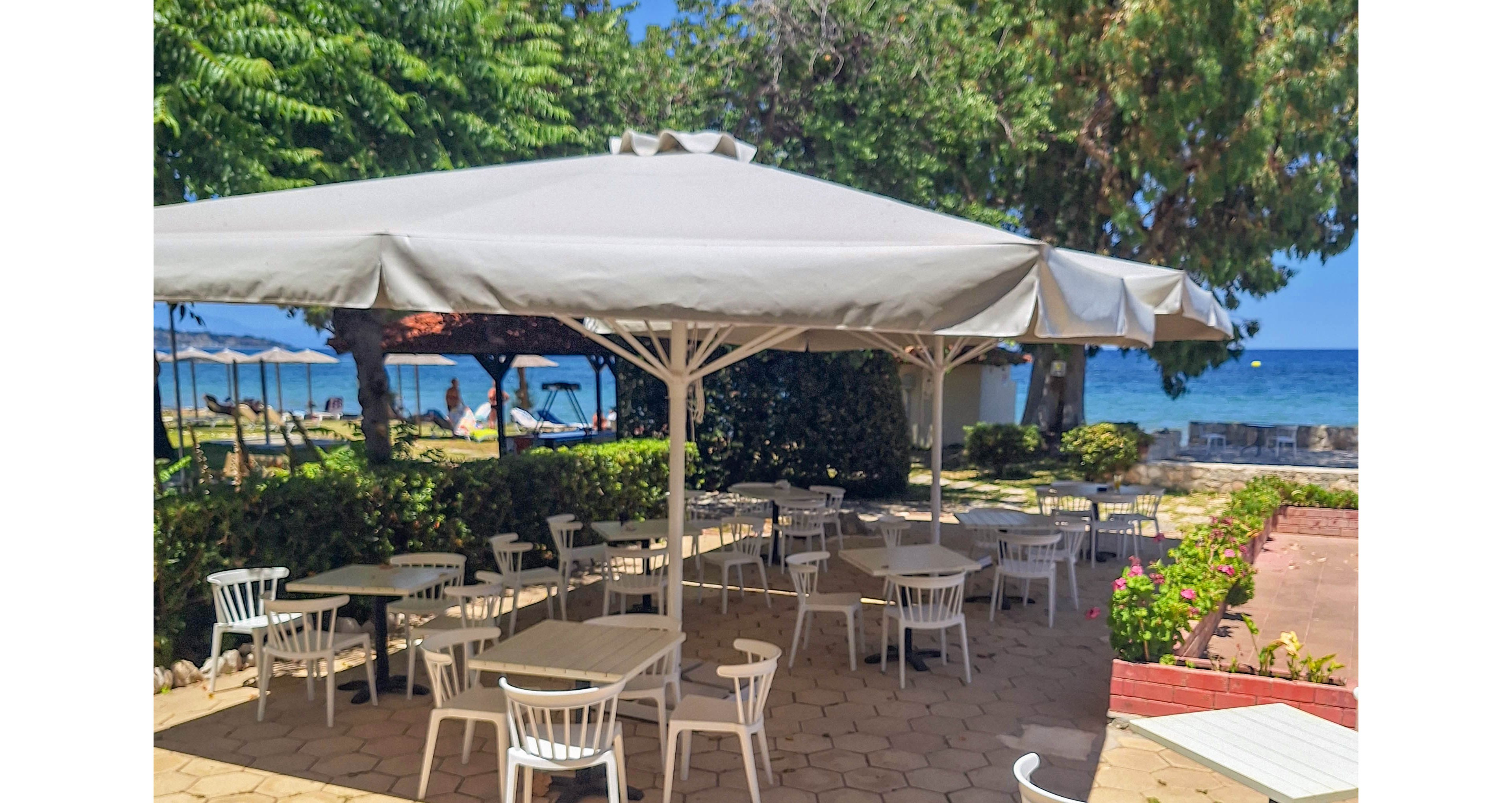 An outdoor café terrace with white tables and chairs sits under large white umbrellas. The seating area is paved with hexagonal tiles and appears quiet and empty. Lush green trees frame the space, leading the eye toward the blue sea in the background. Bright sunlight and clear skies create a relaxed, summery coastal atmosphere.