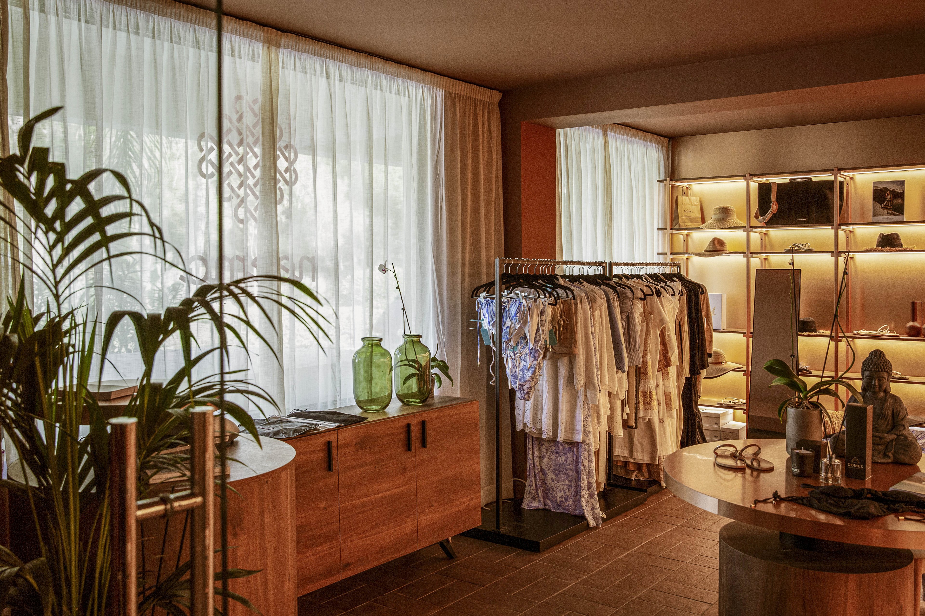 A small boutique interior features a rack of light-colored garments neatly arranged in the center of the room. Soft daylight filters through sheer curtains, illuminating wooden furniture and decorative vases. A backlit shelving unit displays folded textiles and small objects, adding warmth to the space. The overall atmosphere feels calm, inviting, and thoughtfully styled.