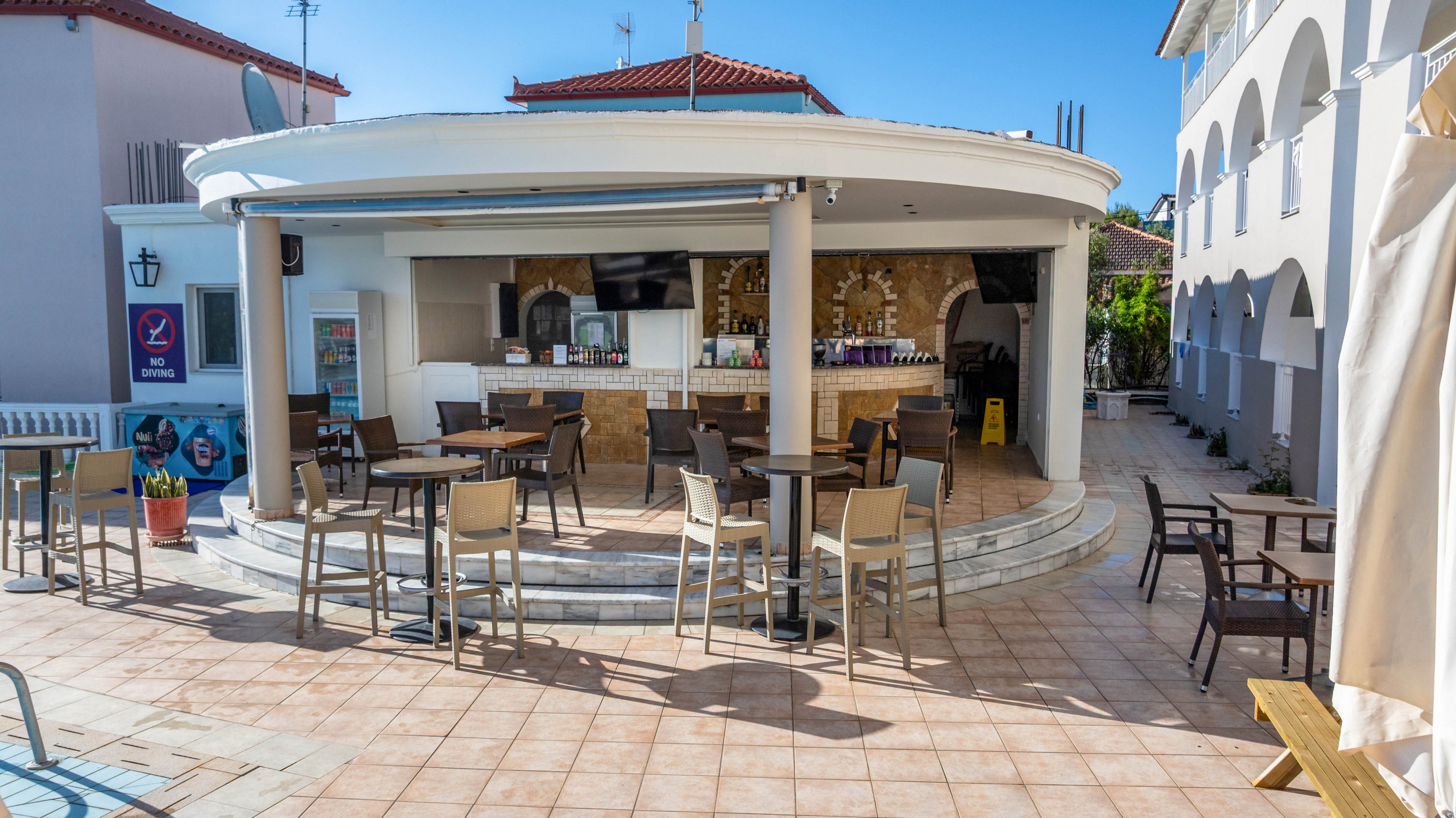An open-air circular bar stands at the center of a tiled patio in a sunny resort setting. Empty tables and chairs are arranged around the bar, suggesting a quiet time between guests. White columns and curved steps frame the bar area, with bottles and glasses visible on the back shelves. Surrounding buildings with arches and red-tiled roofs create a Mediterranean or coastal atmosphere under a clear blue sky.