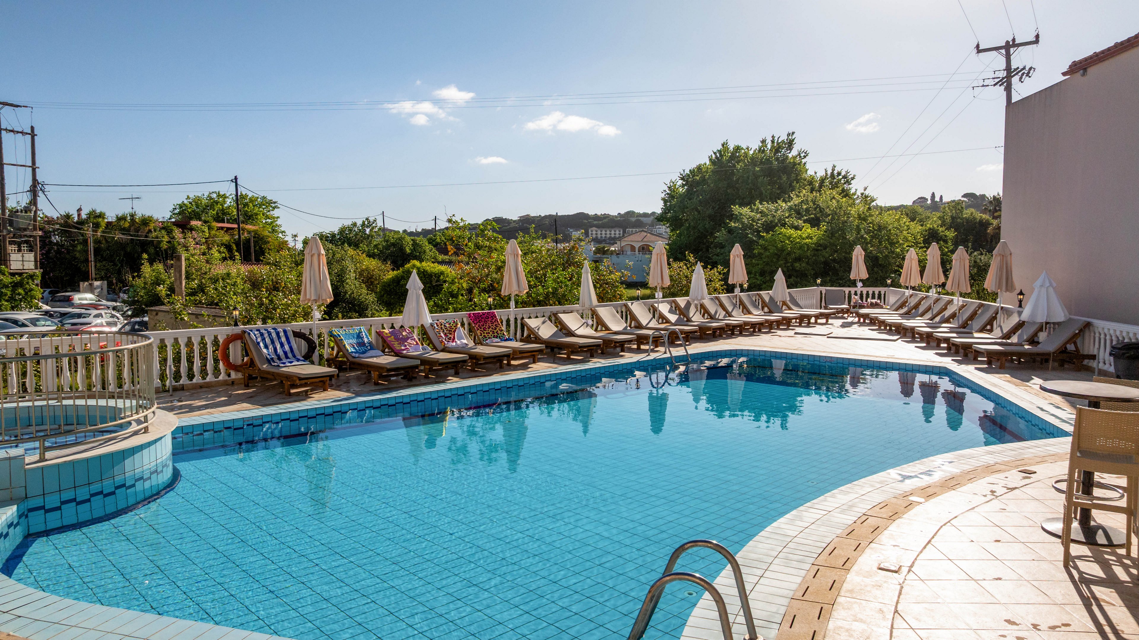 An outdoor hotel swimming pool is surrounded by wooden loungers and closed umbrellas on a sunny day. Colorful towels are draped over the chairs, adding bright accents around the clear blue water. Trees and greenery form a lush backdrop beyond the pool area, with a few buildings and power lines visible in the distance. The scene suggests a quiet, relaxing vacation atmosphere in warm weather.