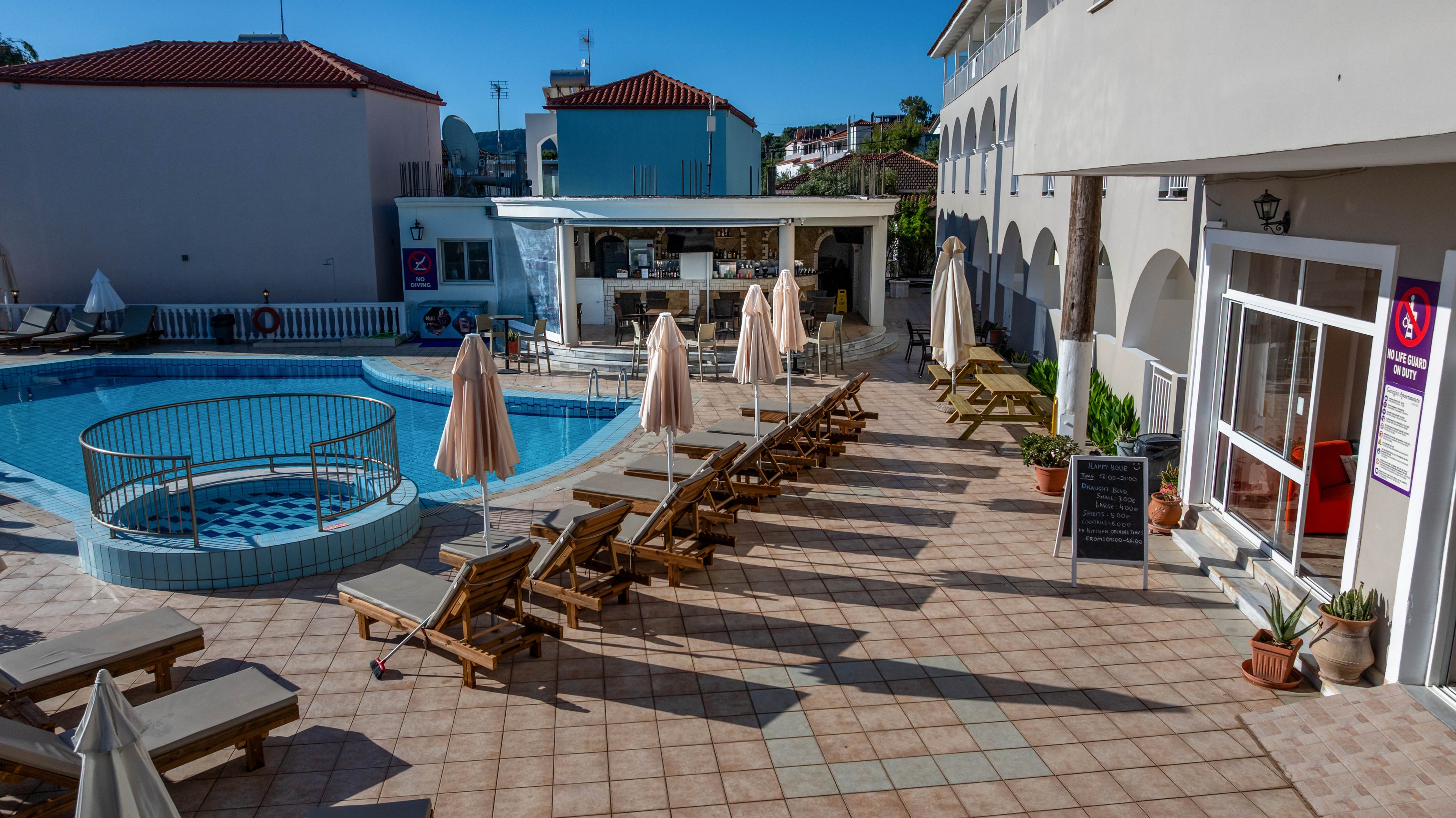 An outdoor hotel pool area is shown under bright daylight with rows of wooden sun loungers and closed umbrellas. A curved swimming pool with a small fenced circular section sits at the center of the tiled terrace. In the background, a pool bar and surrounding low-rise buildings create a relaxed resort atmosphere. The scene appears quiet and empty, suggesting an early morning or off-peak moment at the hotel.