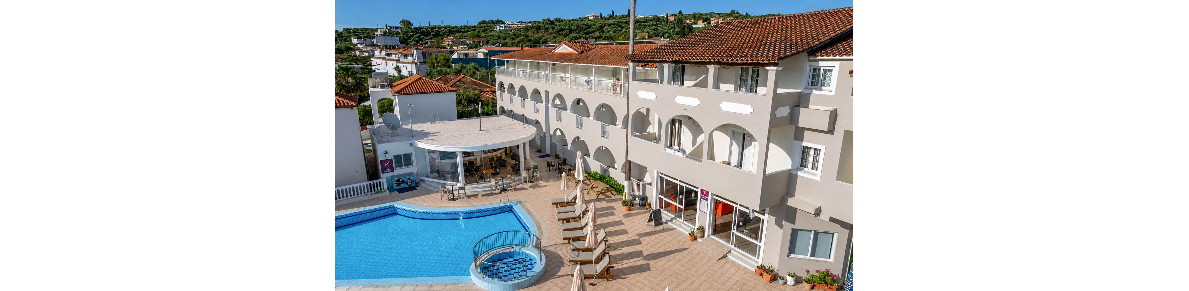 A bright hotel complex with terracotta roofs surrounds a clear blue outdoor swimming pool. Sun loungers and umbrellas are neatly arranged on the tiled poolside terrace, creating a relaxed vacation atmosphere. A circular pool bar structure sits beside the main pool, with tables and chairs under its canopy. The upper floors feature balconies overlooking the pool area and the green hillside landscape in the background.