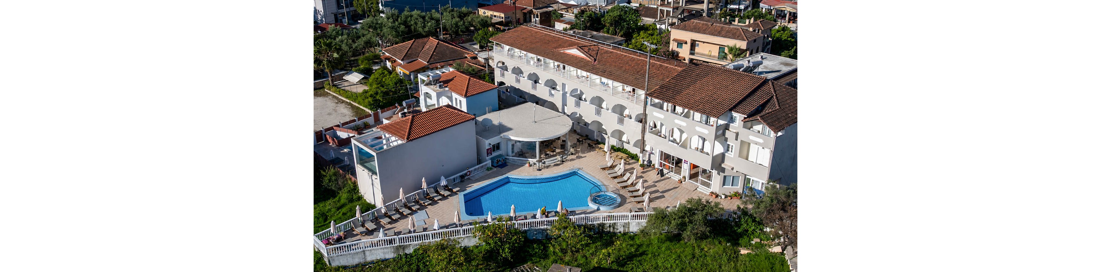 A large white hotel complex with red-tiled roofs is shown from an aerial perspective. At the center of the property is a blue outdoor swimming pool surrounded by sun loungers and umbrellas. The building features multiple balconies and arched openings facing the pool terrace. Green vegetation and neighboring houses frame the resort, suggesting a warm Mediterranean or coastal town setting.