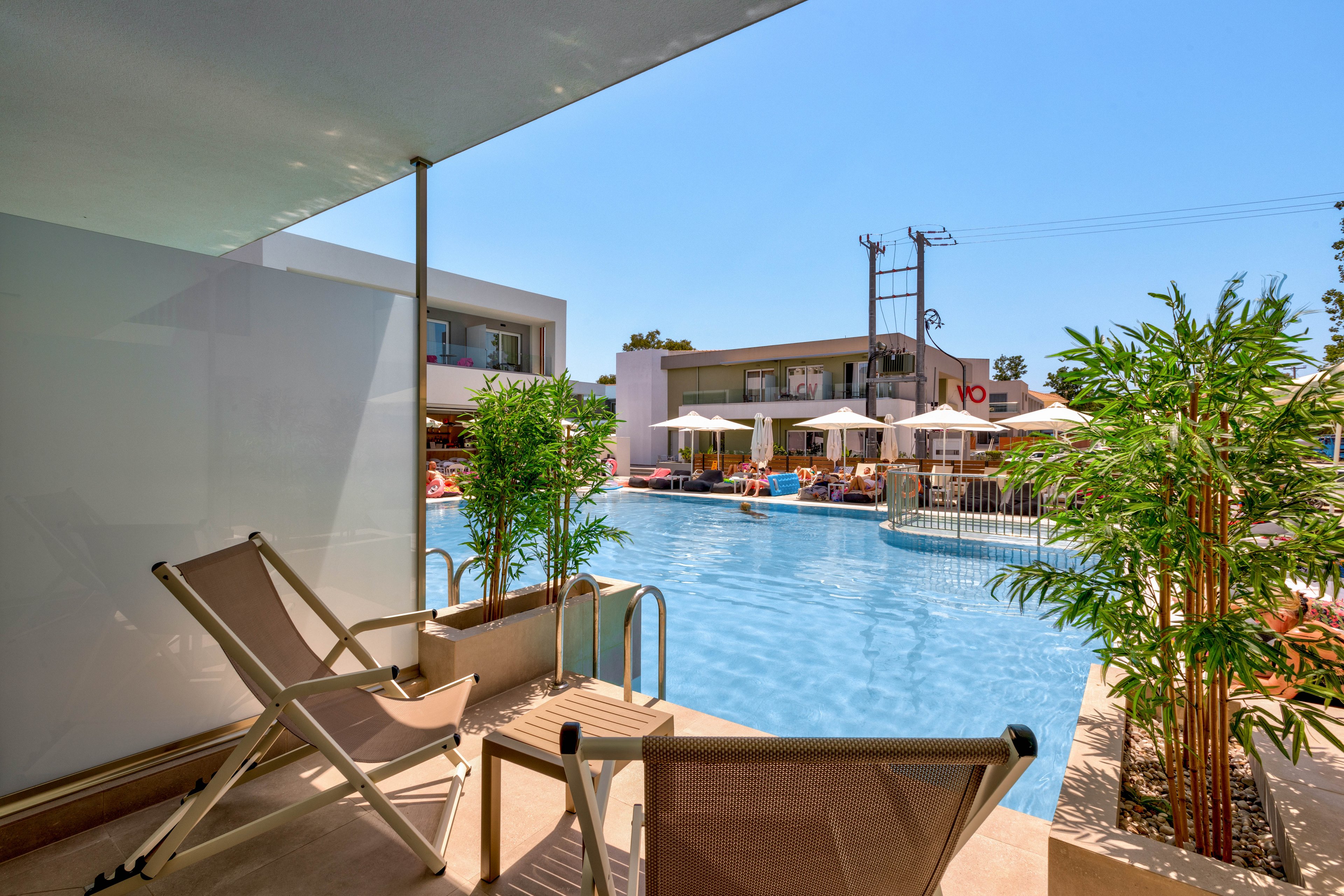 A modern hotel terrace opens directly onto a bright outdoor swimming pool. Two reclining lounge chairs sit on the shaded patio beside a metal pool ladder. Across the pool, guests relax under white umbrellas near a bar or café area. Contemporary low-rise buildings and clear blue sky create a sunny resort atmosphere. Green potted plants add a touch of nature to the sleek architectural setting.
