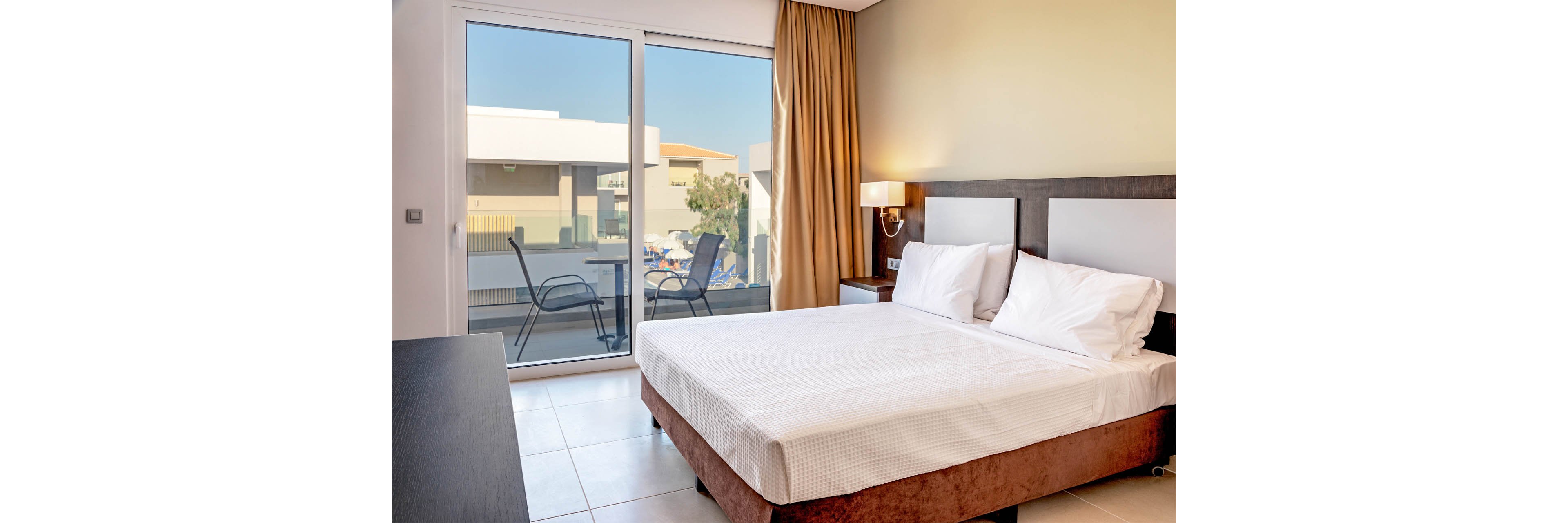 A modern hotel bedroom features a neatly made double bed with white linens and pillows. Large sliding glass doors open onto a balcony with two chairs and a small table. Warm beige walls, a bedside lamp, and floor-length curtains create a cozy yet contemporary atmosphere. Outside, neighboring buildings and clear blue sky are visible, suggesting a sunny vacation destination.