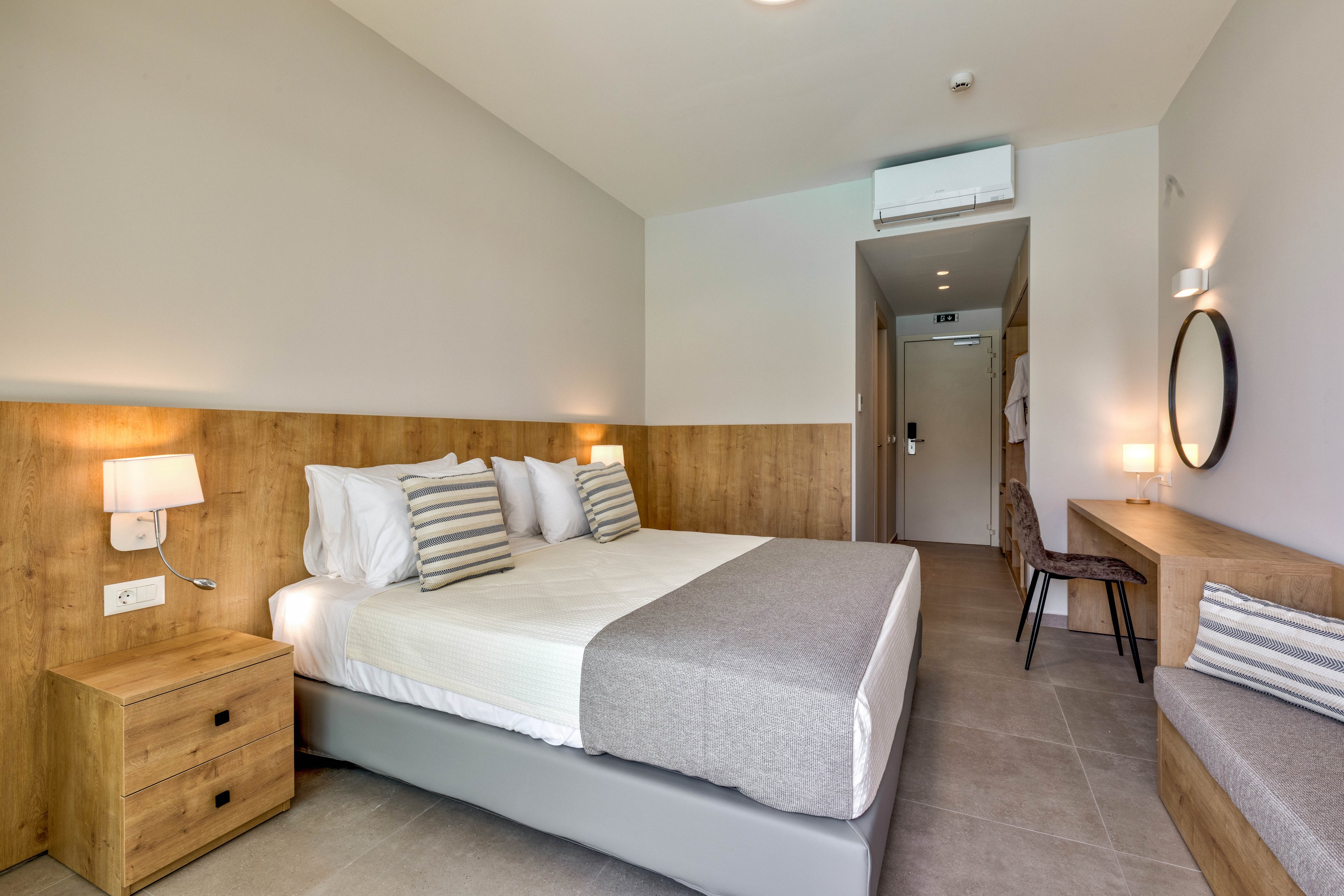A contemporary hotel room features a neatly made double bed with white linens and gray accents. The space includes wooden wall paneling, a bedside table, and built-in lighting on both sides of the bed. A desk with a chair, a round wall mirror, and a cushioned bench line the opposite wall. Neutral tones and clean lines create a calm, minimalist atmosphere in the bright interior.
