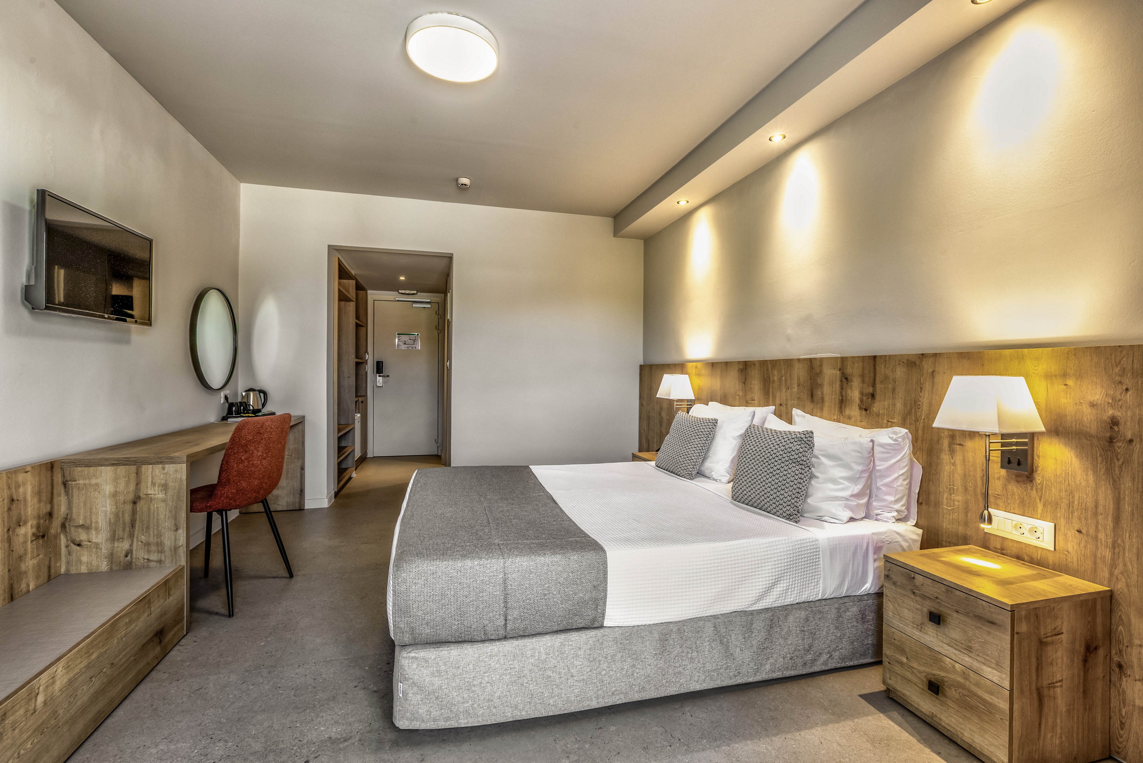 A contemporary hotel room features a neatly made double bed with gray and white bedding and accent pillows. Warm wooden furniture, including a bedside table and long desk, adds a cozy touch to the minimalist design. A wall-mounted television, round mirror, and desk chair create a functional workspace. Soft lighting from ceiling fixtures and bedside lamps brightens the neutral-toned interior, leading toward the entrance door and hallway.