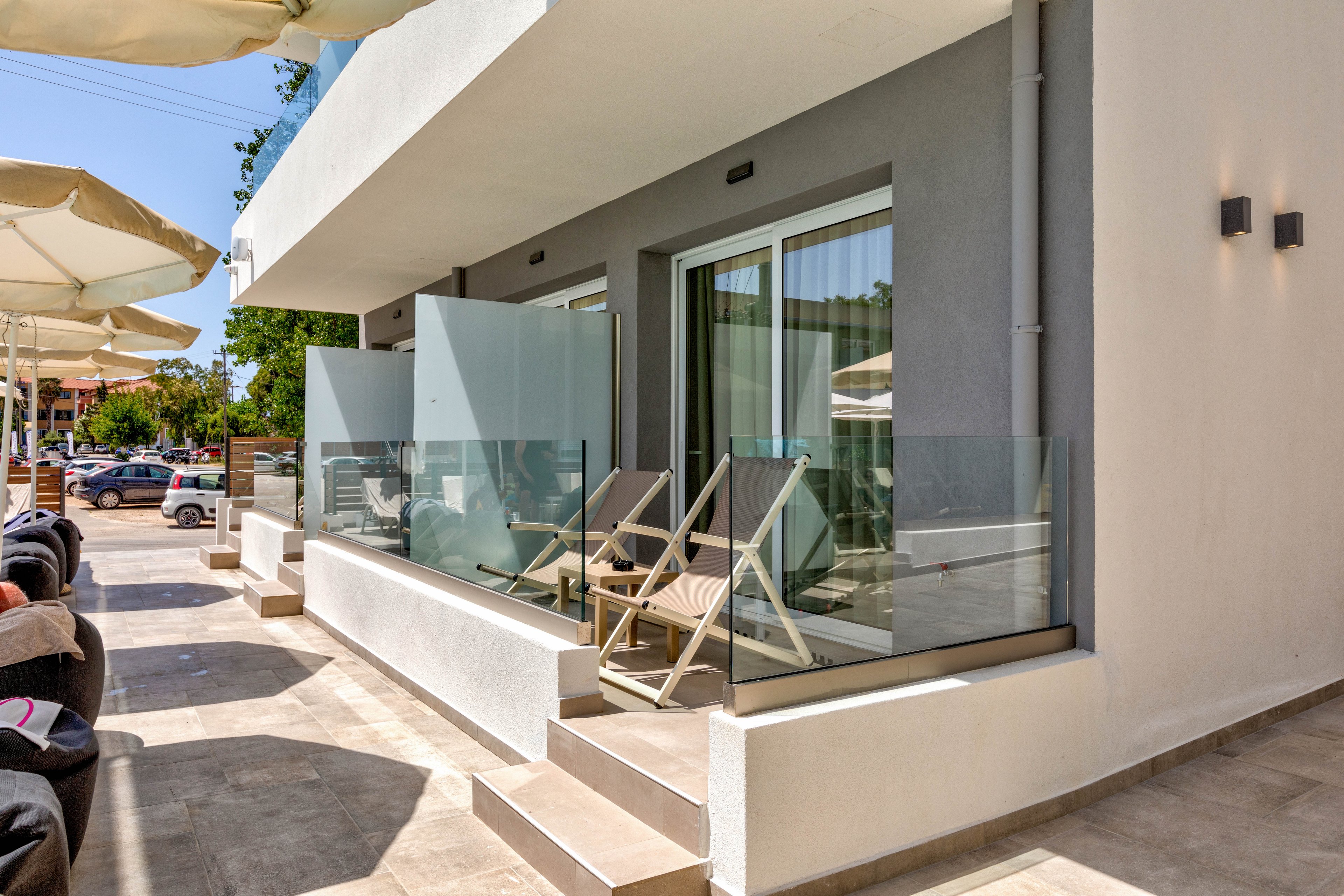 Exterior view of a modern hotel or apartment terrace featuring private patios with glass railings. Two wooden lounge chairs sit on each small balcony in front of large sliding glass doors with curtains. The space is paved with light tiles and shaded by large outdoor umbrellas. Parked cars and greenery are visible in the background, suggesting a sunny vacation destination.