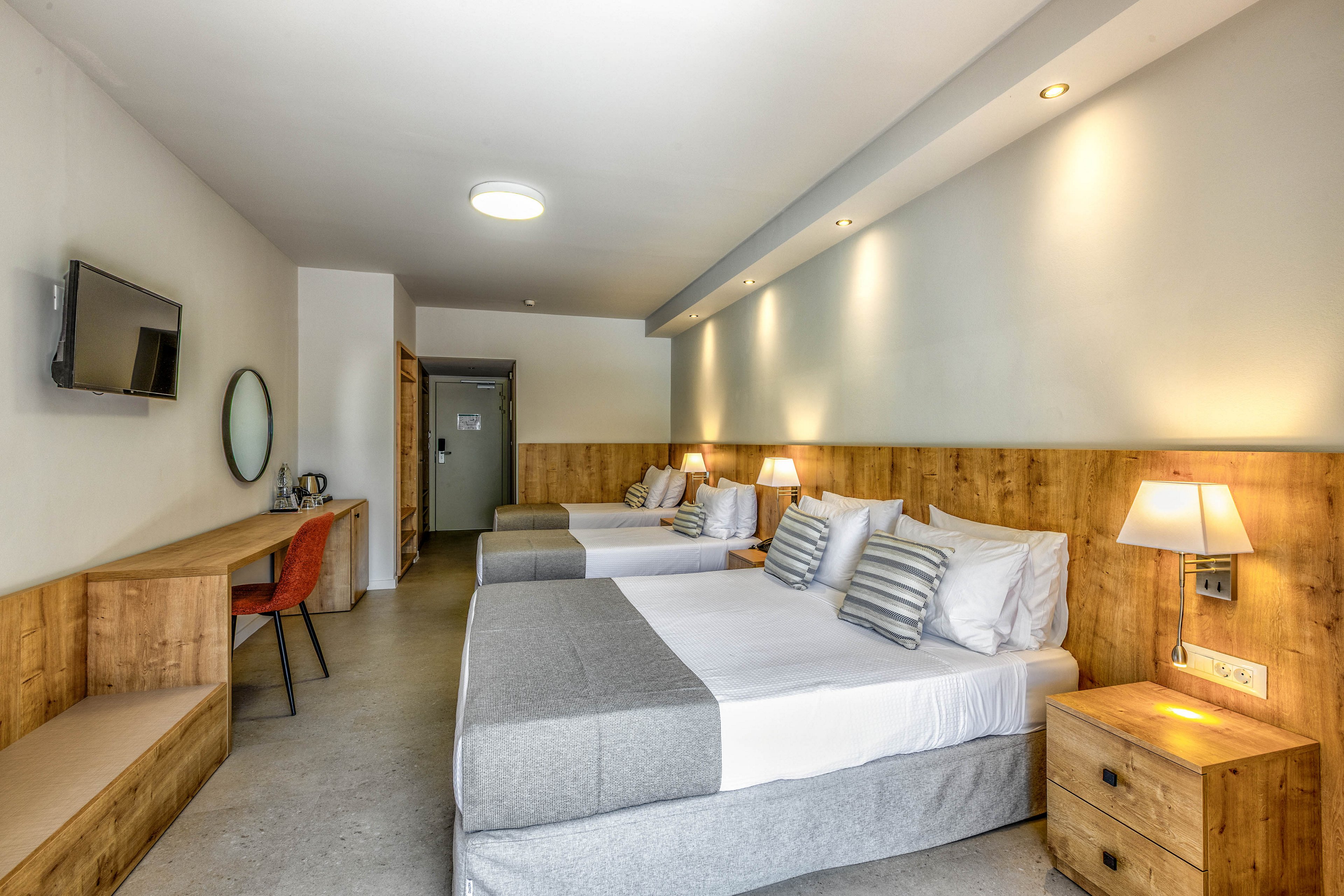 Contemporary hotel room interior featuring three neatly made beds with gray and white bedding. The space includes wooden headboards, bedside tables, and warm wall-mounted lamps creating a cozy atmosphere. A flat-screen TV is mounted on the wall opposite a long wooden desk with an orange chair and round mirror. Neutral colors and clean lines give the room a minimalist, modern feel suitable for families or groups.