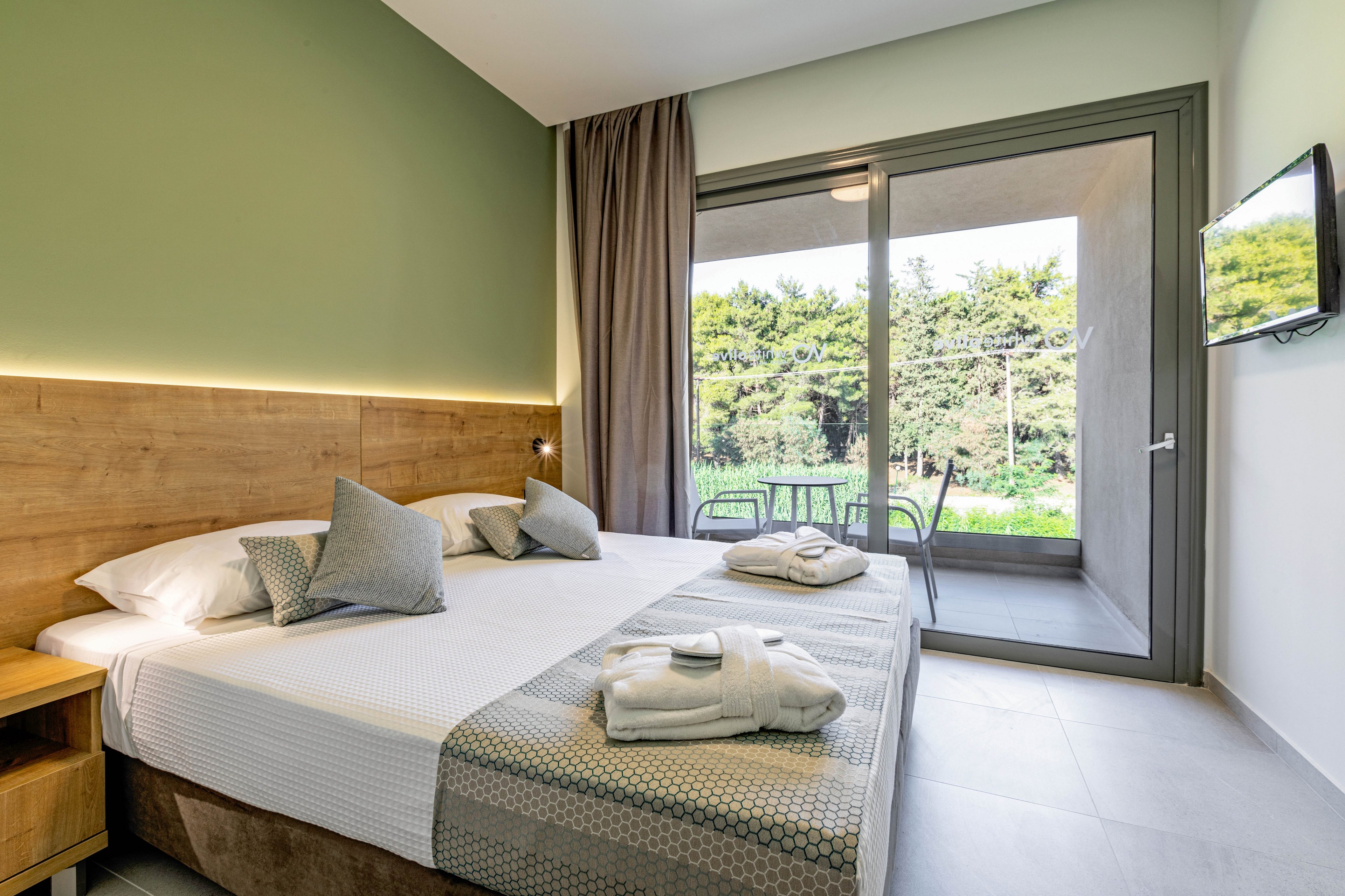 Contemporary hotel room featuring a large double bed neatly arranged with pillows and folded towels. The space opens onto a small terrace with chairs and a table, overlooking a lush green garden. Soft neutral tones, wood accents, and indirect lighting create a calm, inviting atmosphere. A wall-mounted television and floor-to-ceiling glass doors add to the modern, comfortable design.