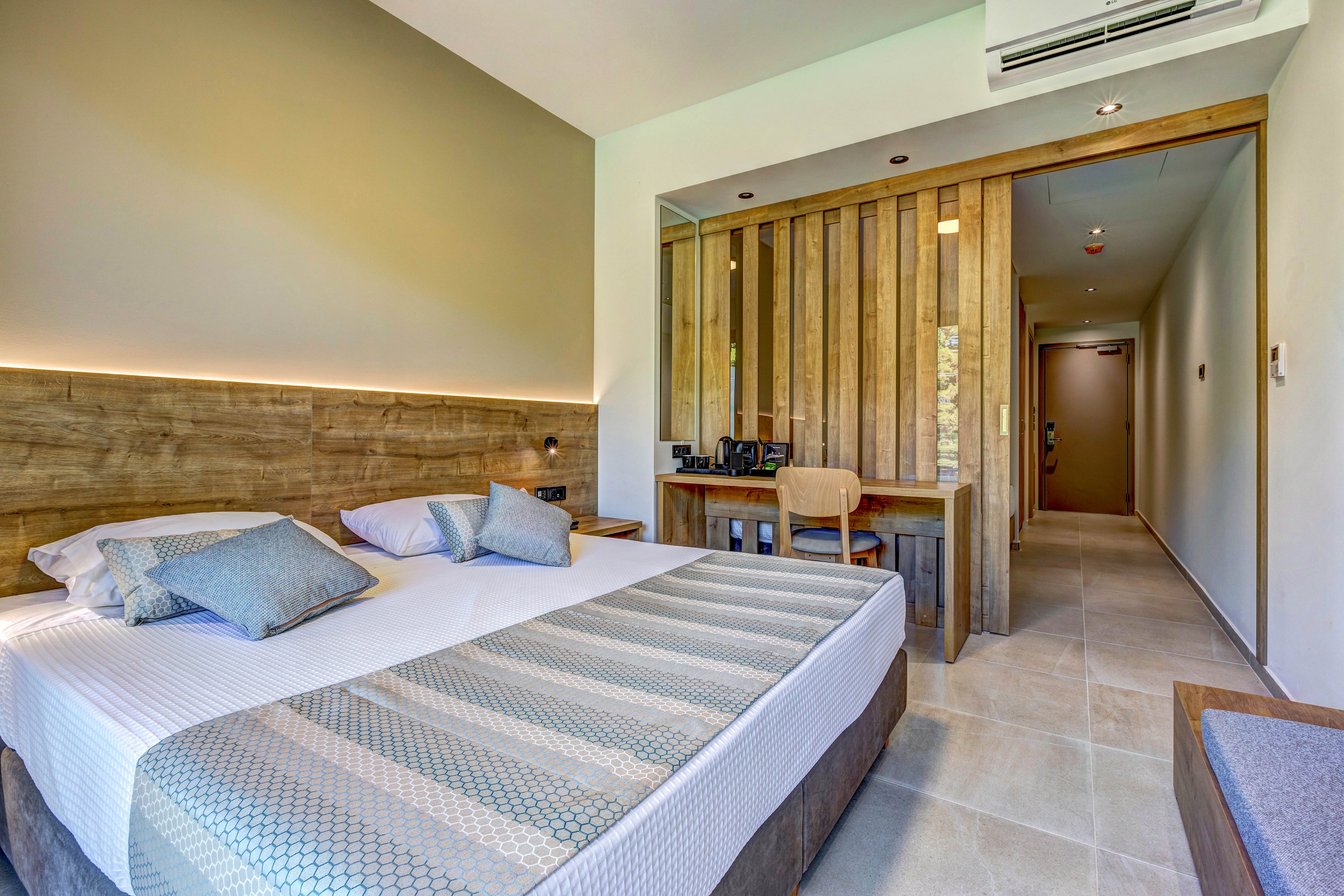 Contemporary hotel room featuring a large double bed with patterned bedding and multiple pillows. The space includes a wooden headboard with integrated lighting and a work desk area separated by vertical wooden slats. Neutral tones and soft lighting create a calm, minimalist atmosphere. A hallway leading to the entrance door and tiled flooring complete the clean, modern design.